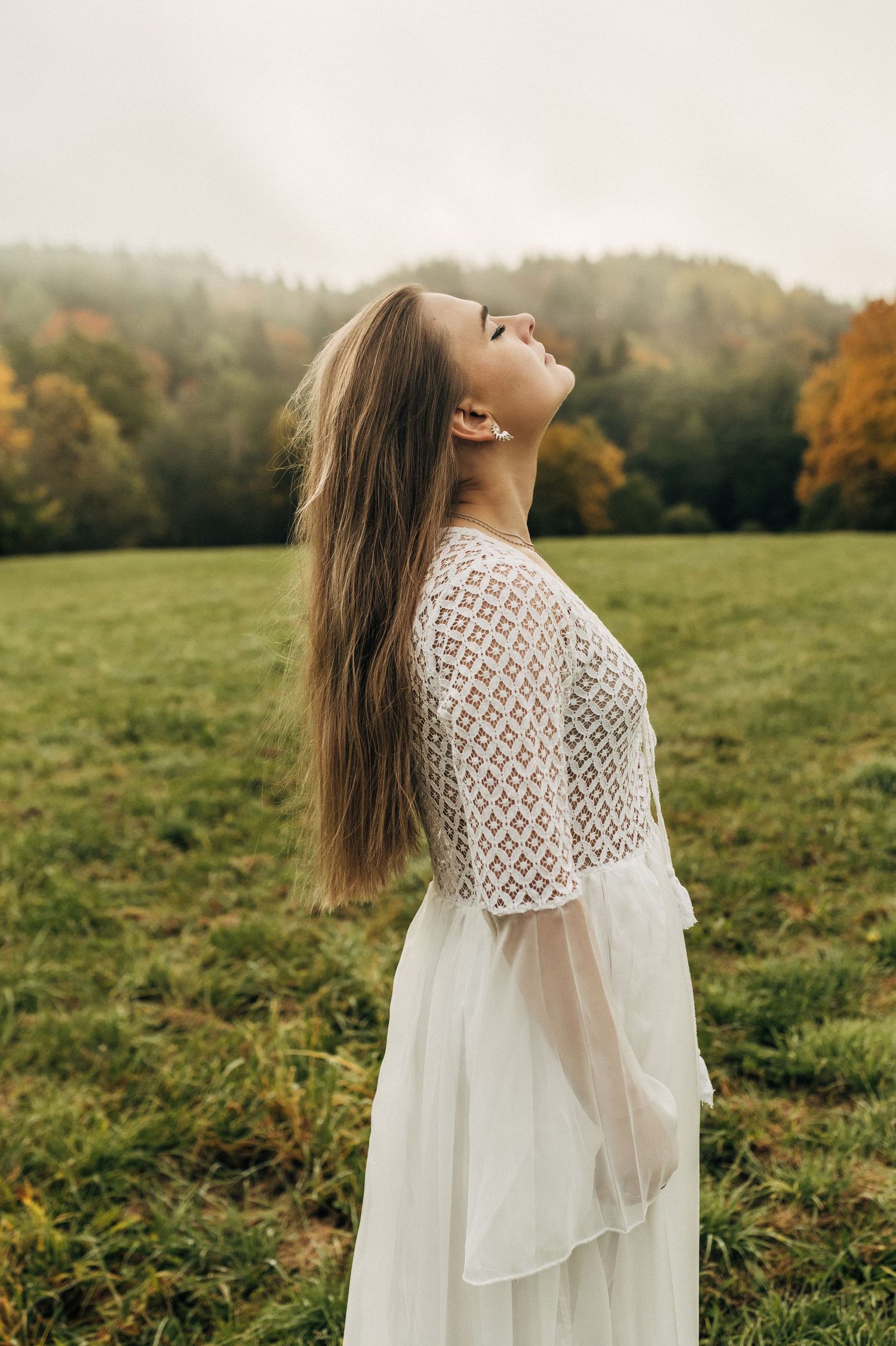 Woman in flowing white lace dress stands in autumn meadow with golden light and trees in background.