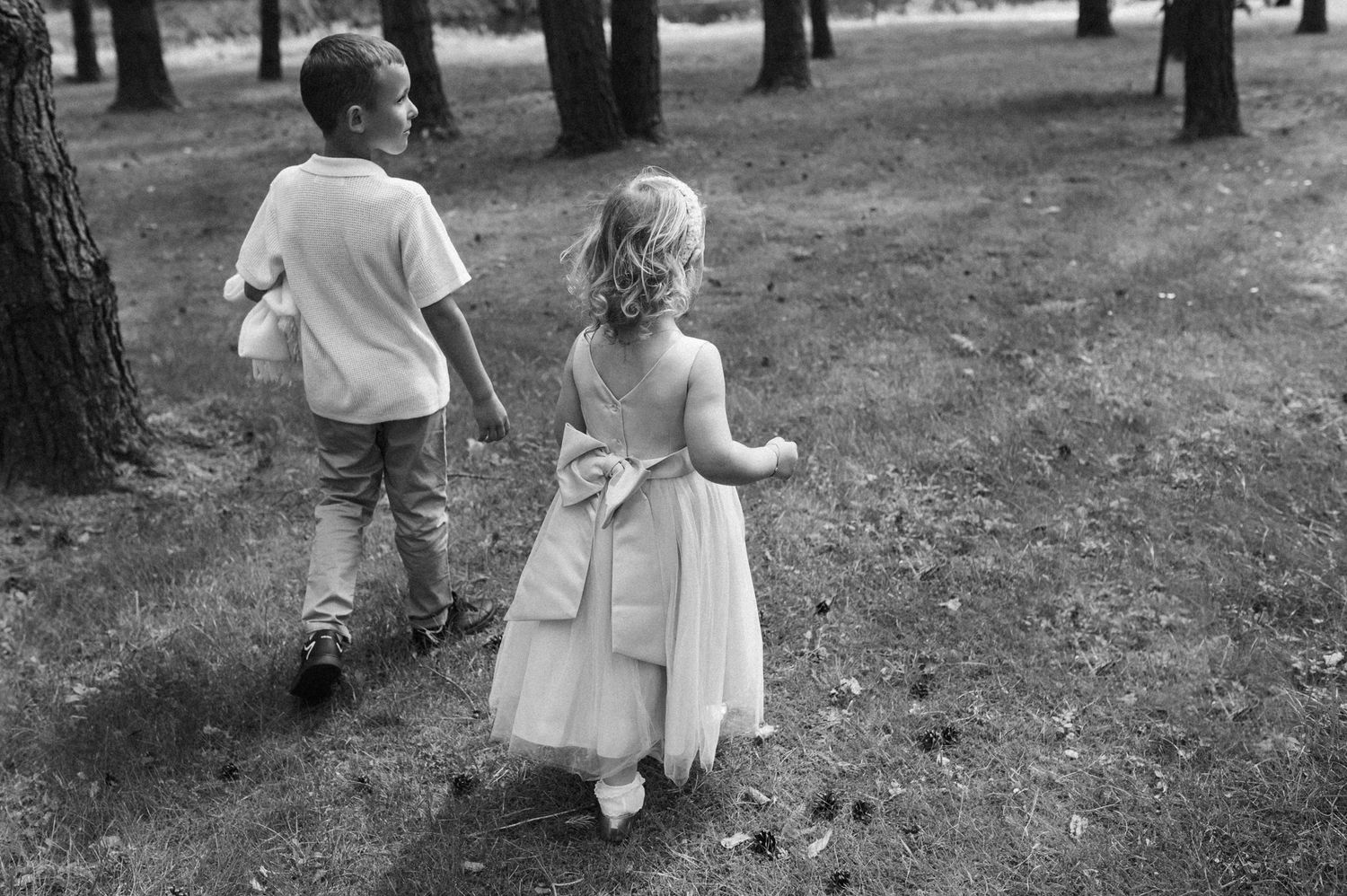 Boy and girl in formal wear walking hand in hand through a forest in black and white.