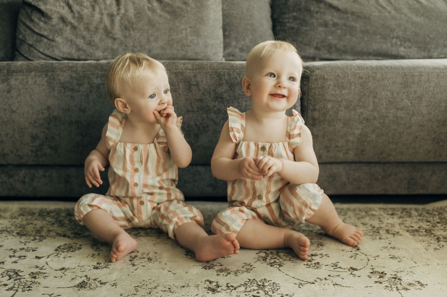 Two babies sitting on a grey floor giggling and laughing together while wearing matching outfits.