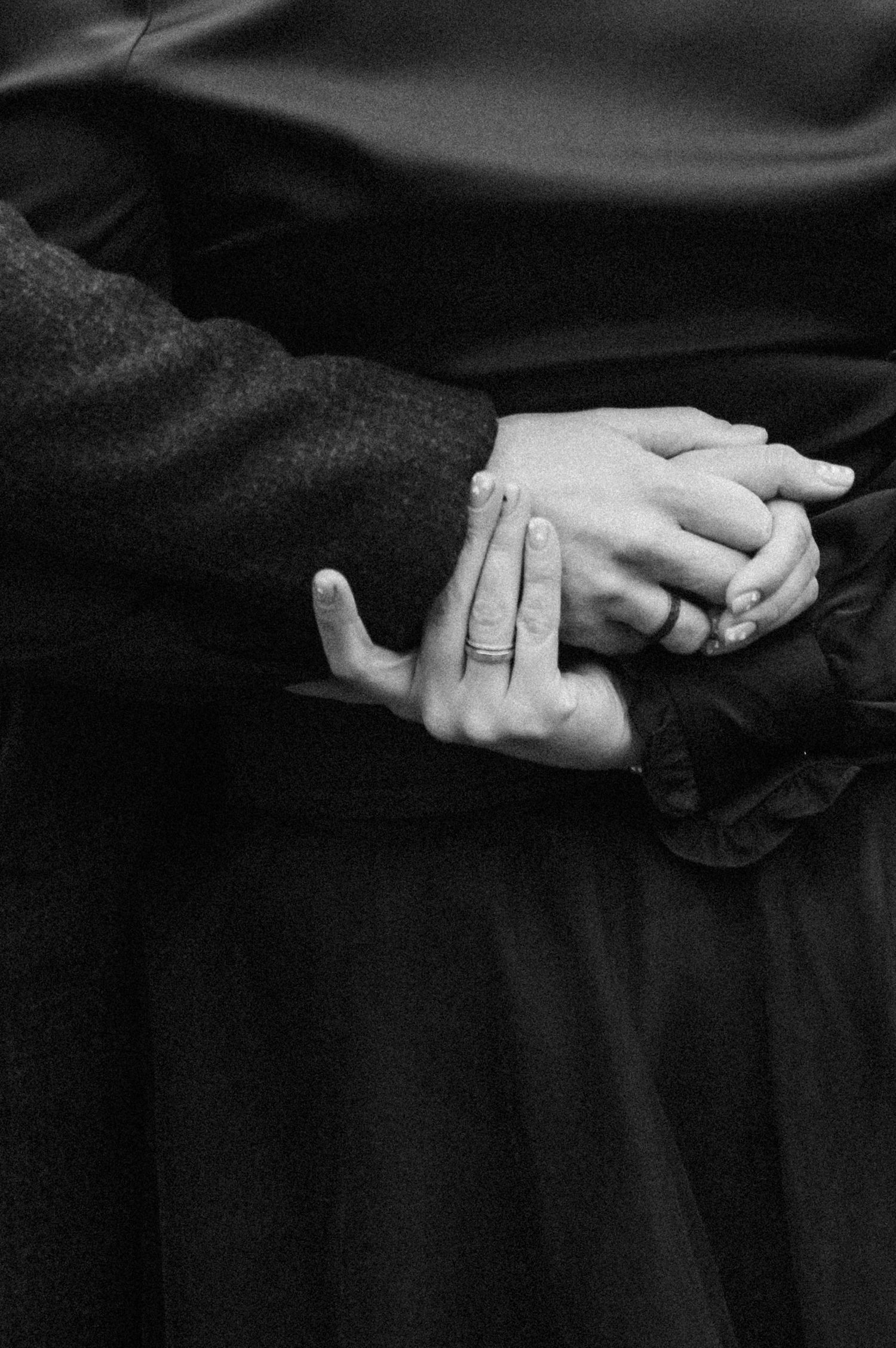 Black and white close-up of folded hands with rings against dark fabric.