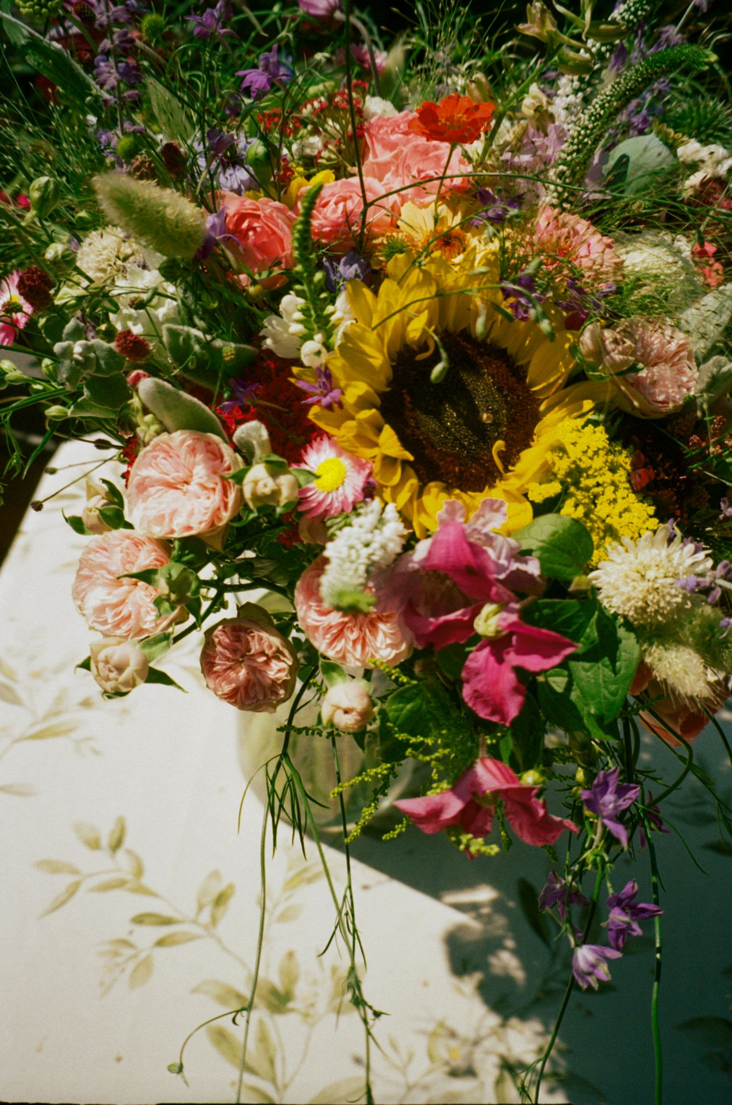 Vibrant mixed flower bouquet with sunflower, pink roses, and wildflowers arranged on a floral-patterned surface.