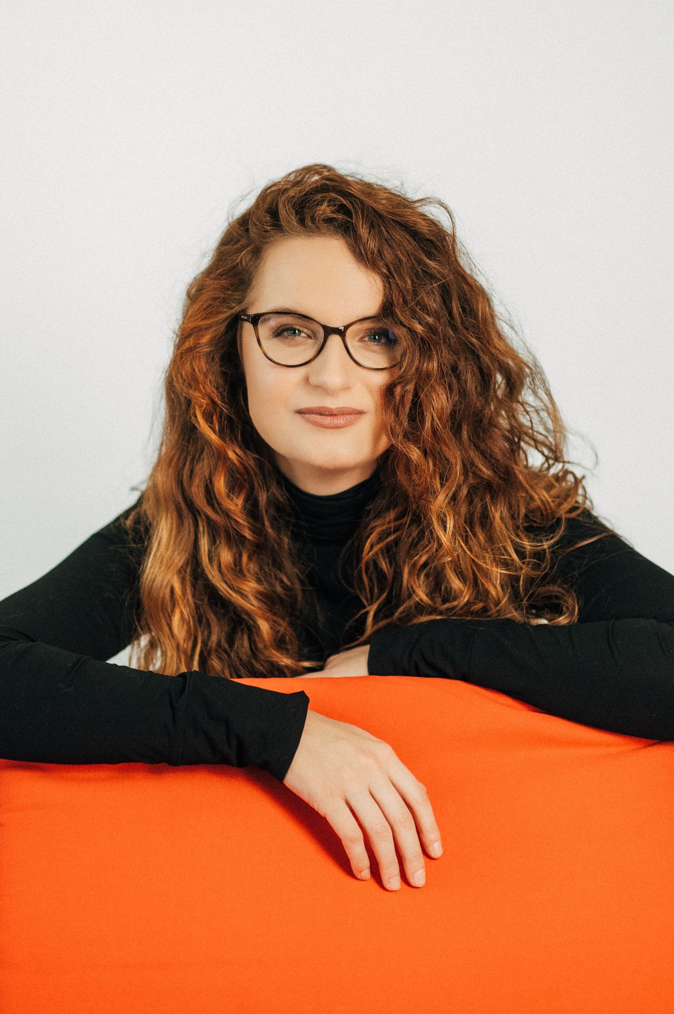 Portrait with curly hair and glasses leaning on bright orange surface.