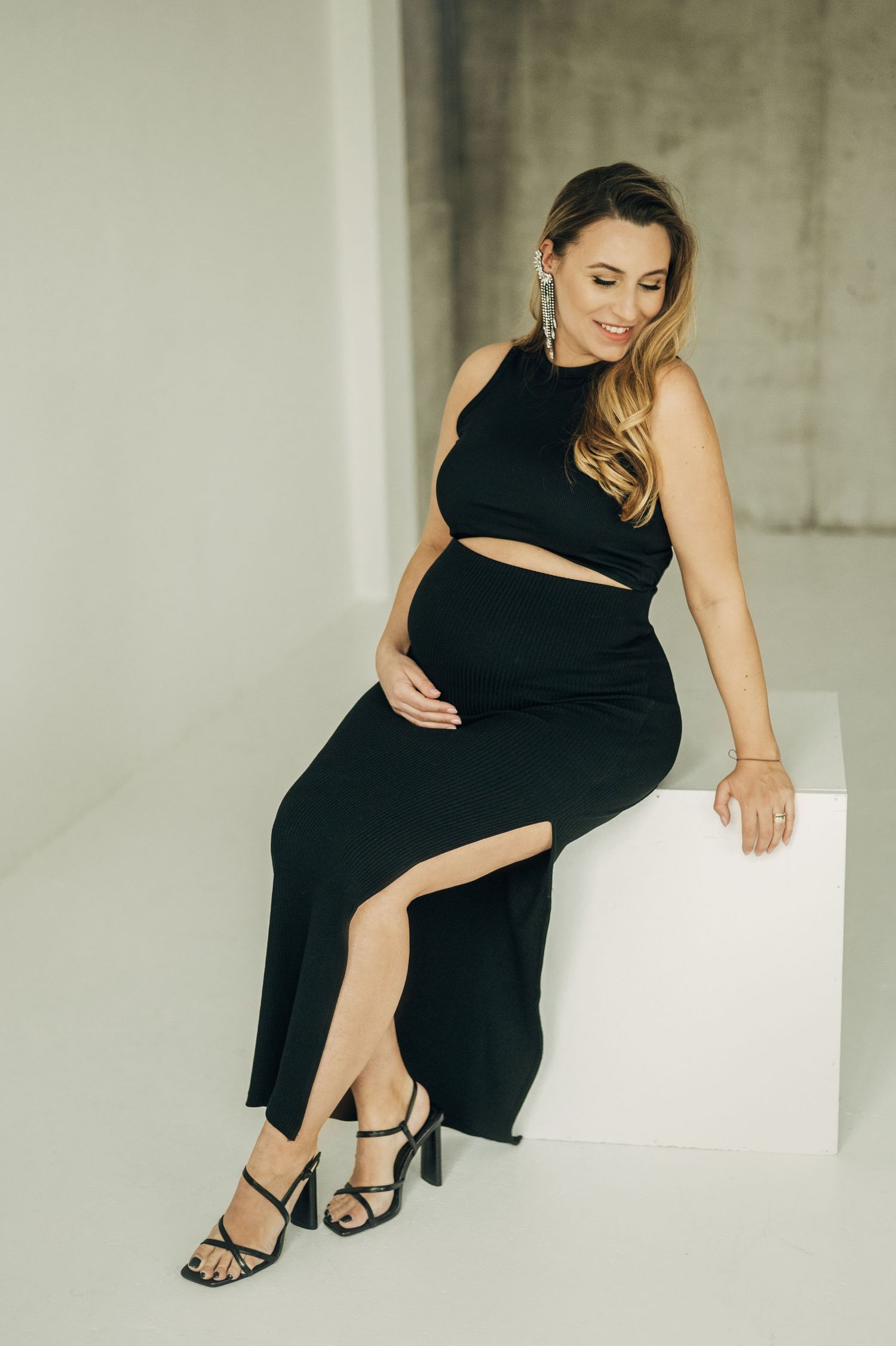 Stylish maternity session with woman in black dress posing on white furniture.