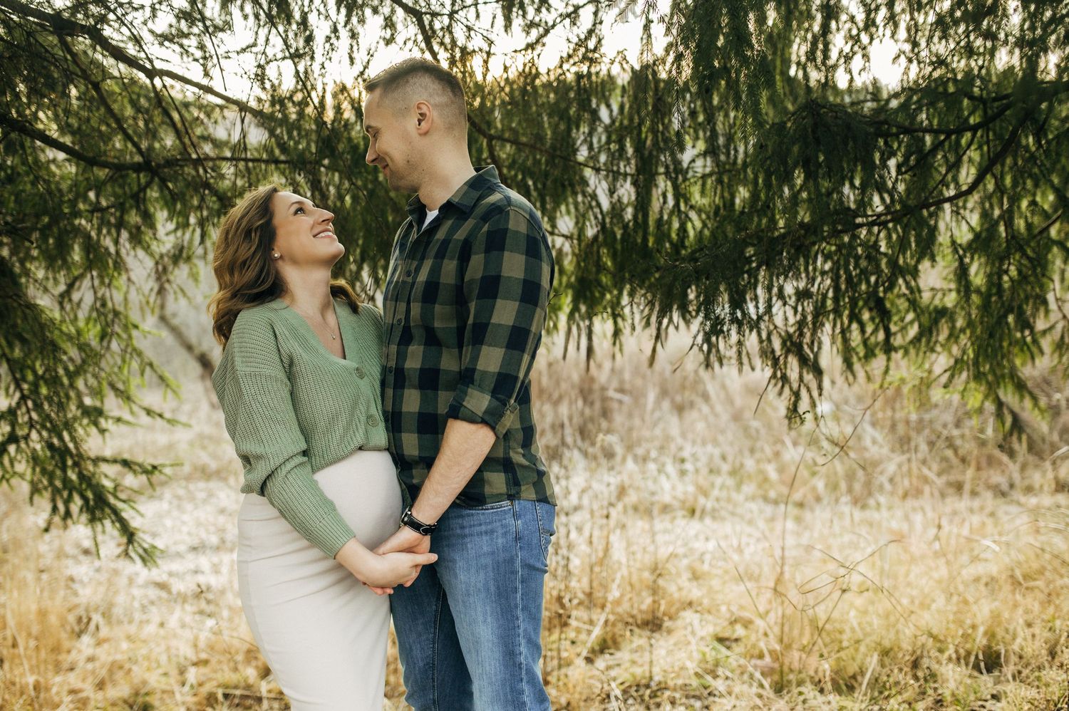Happy couple embracing outdoors under pine branches, woman in green sweater showing baby bump, man in plaid shirt.