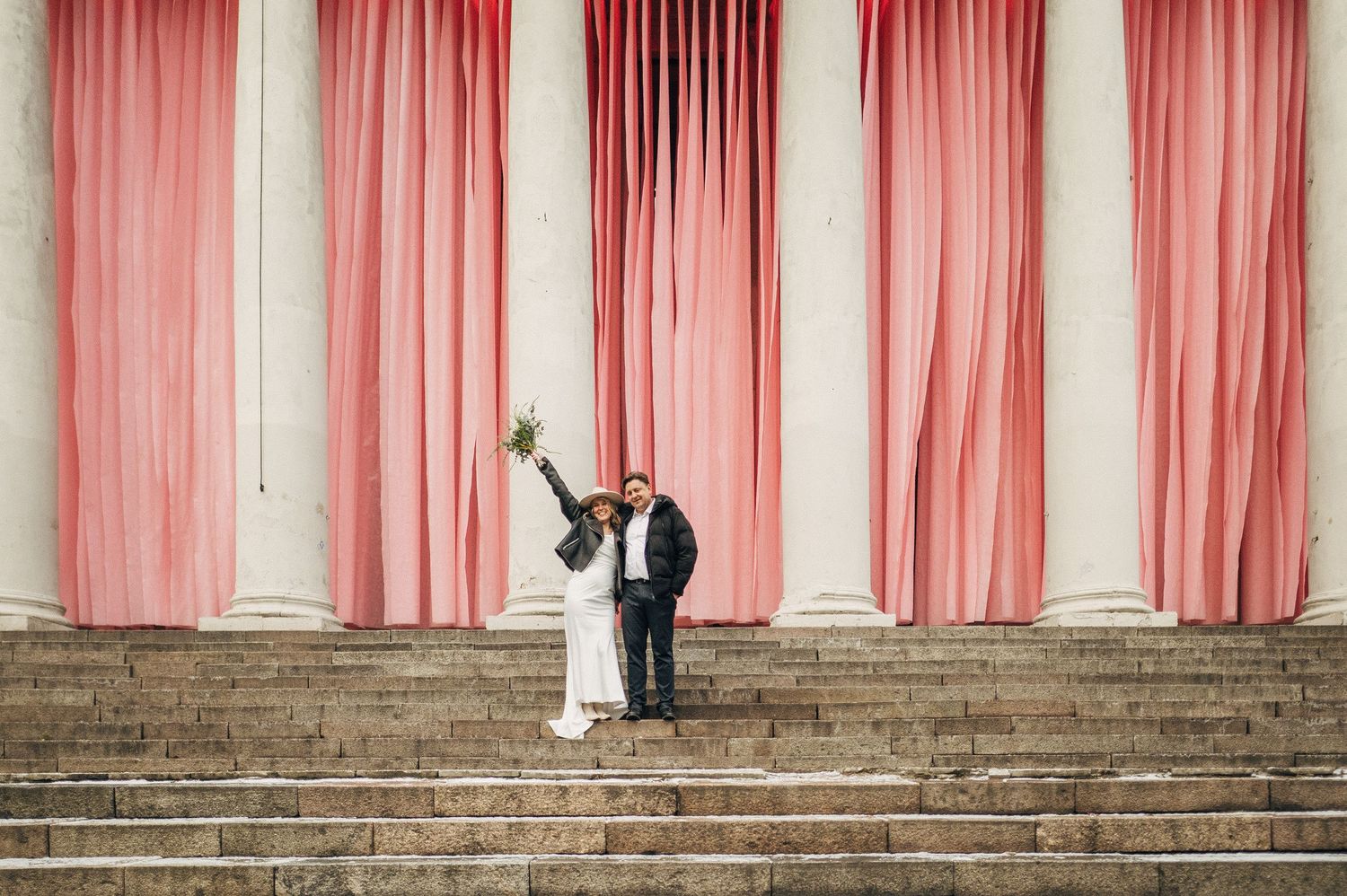 A wedding couple poses on grand white stone steps with pink curtains creating an elegant backdrop for their portraits.