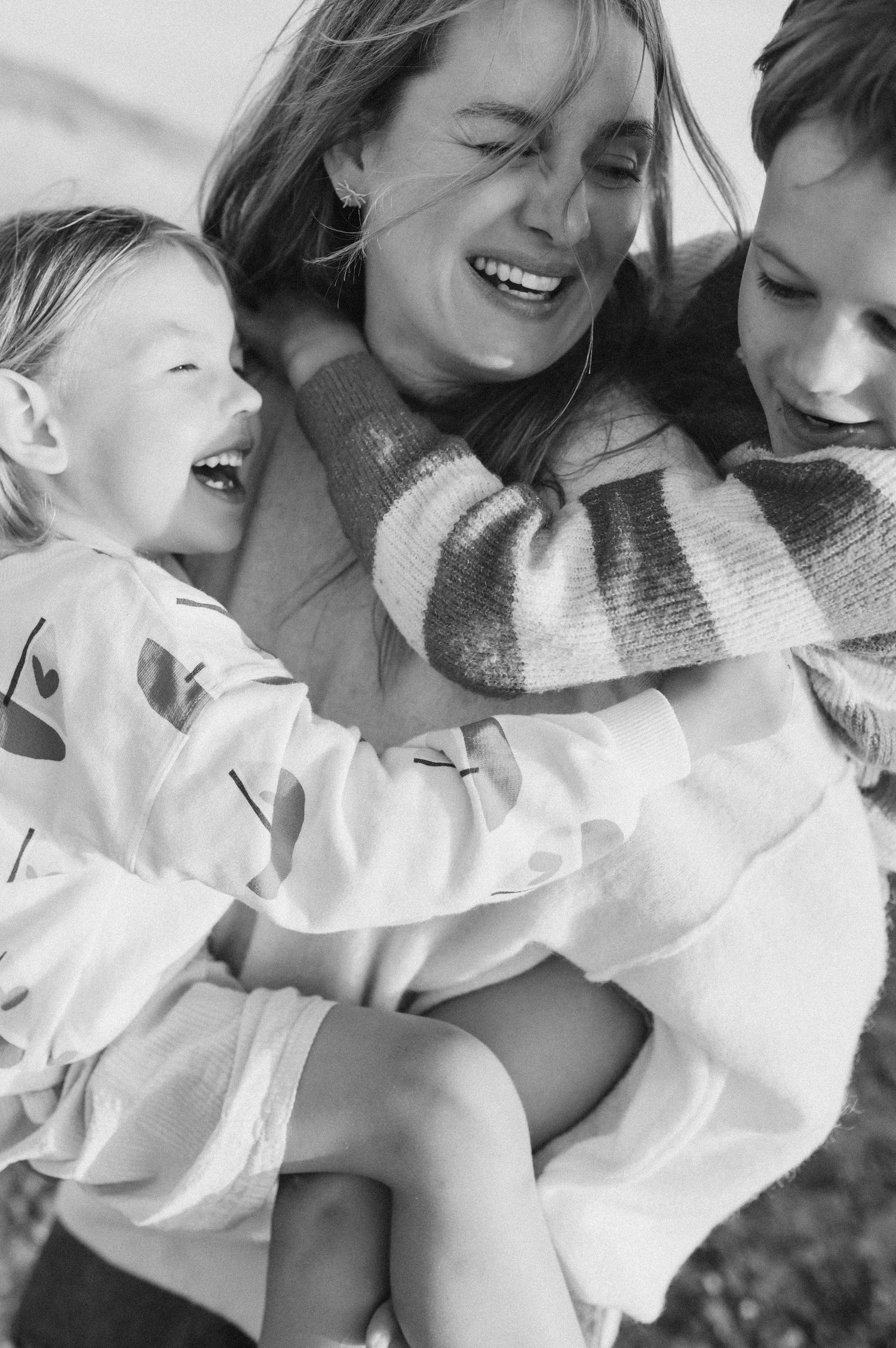 Black and white photo of a laughing mother hugging two happy children outdoors.