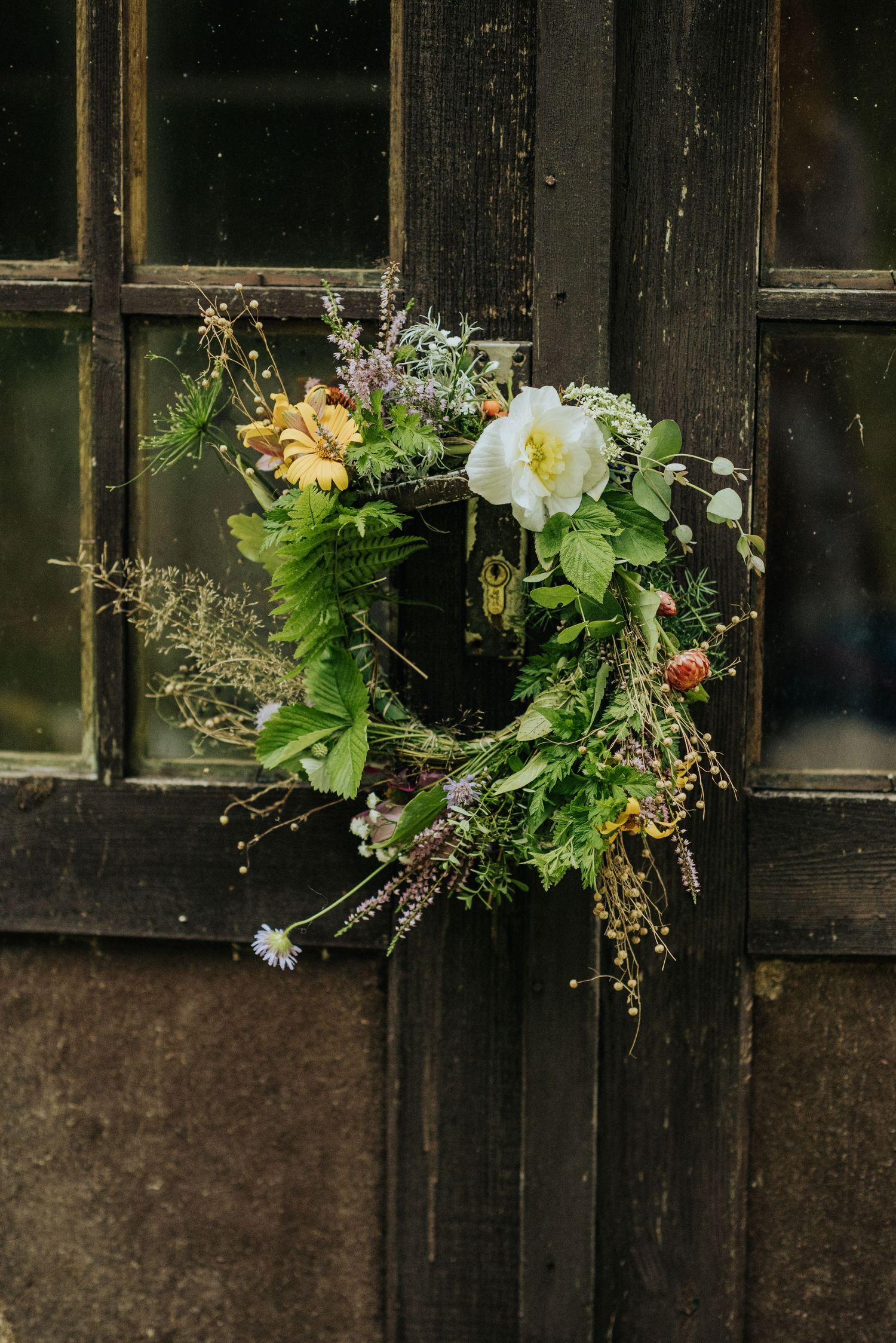 A rustic wildflower wreath with white and yellow blooms hanging on a dark wooden door.