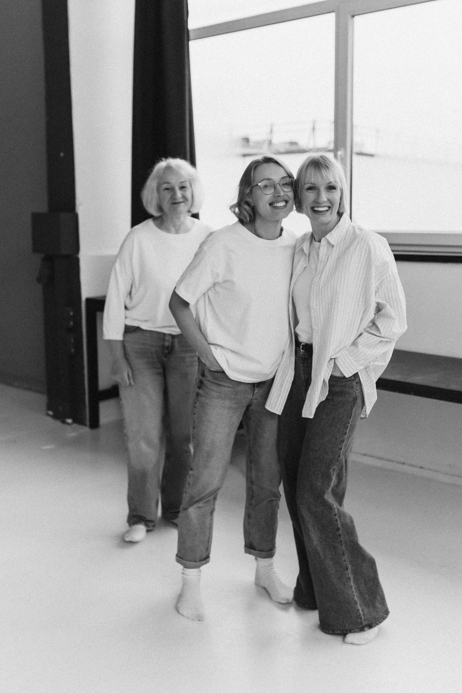 Three people in matching white shirts and jeans lean together laughing in a bright studio setting.
