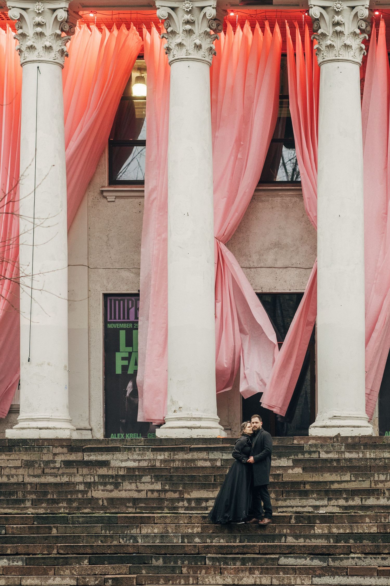 Couple on stone steps beneath coral-colored draped columns.