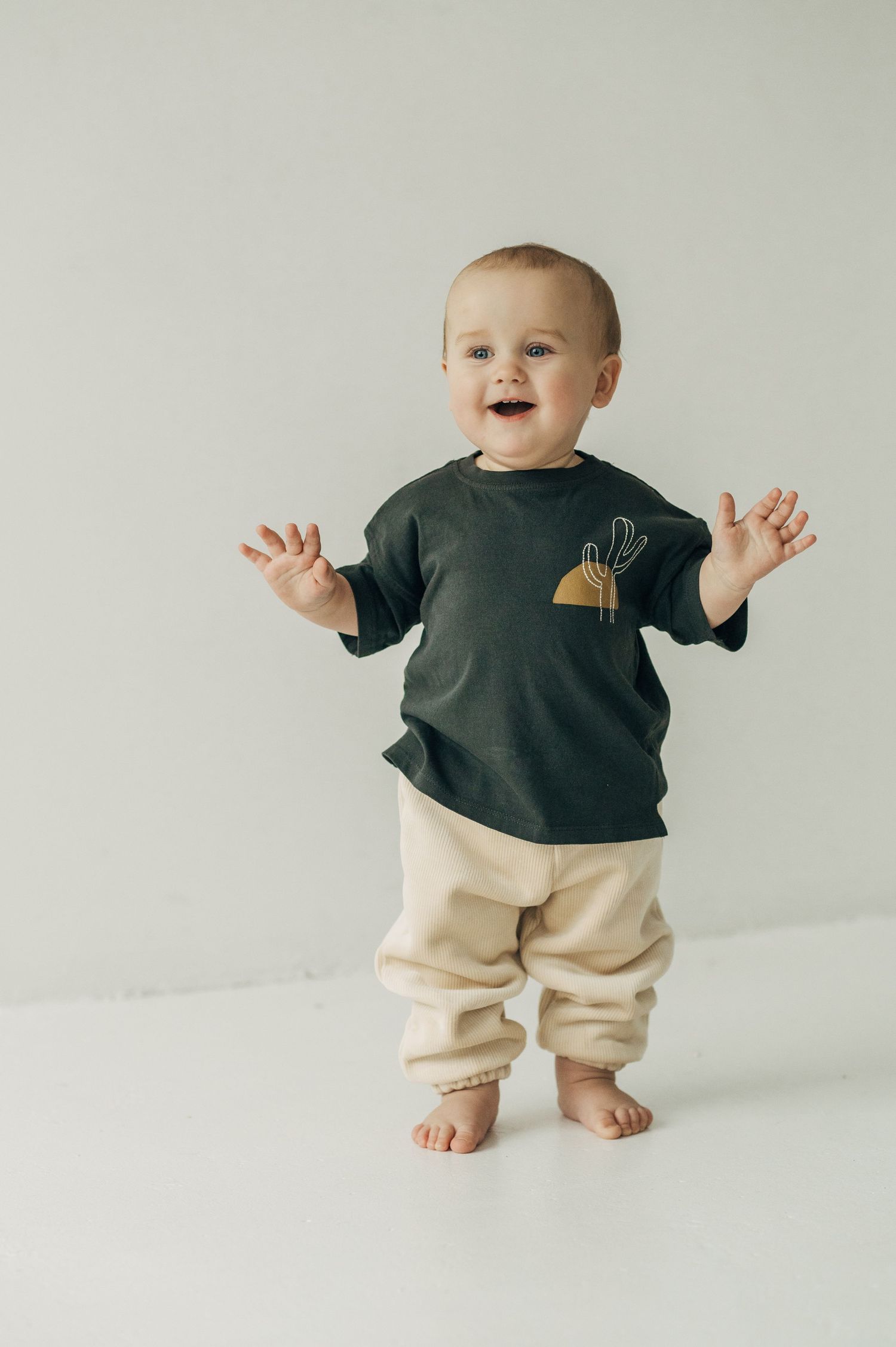 Baby wearing black shirt and cream pants standing and balancing against white background.