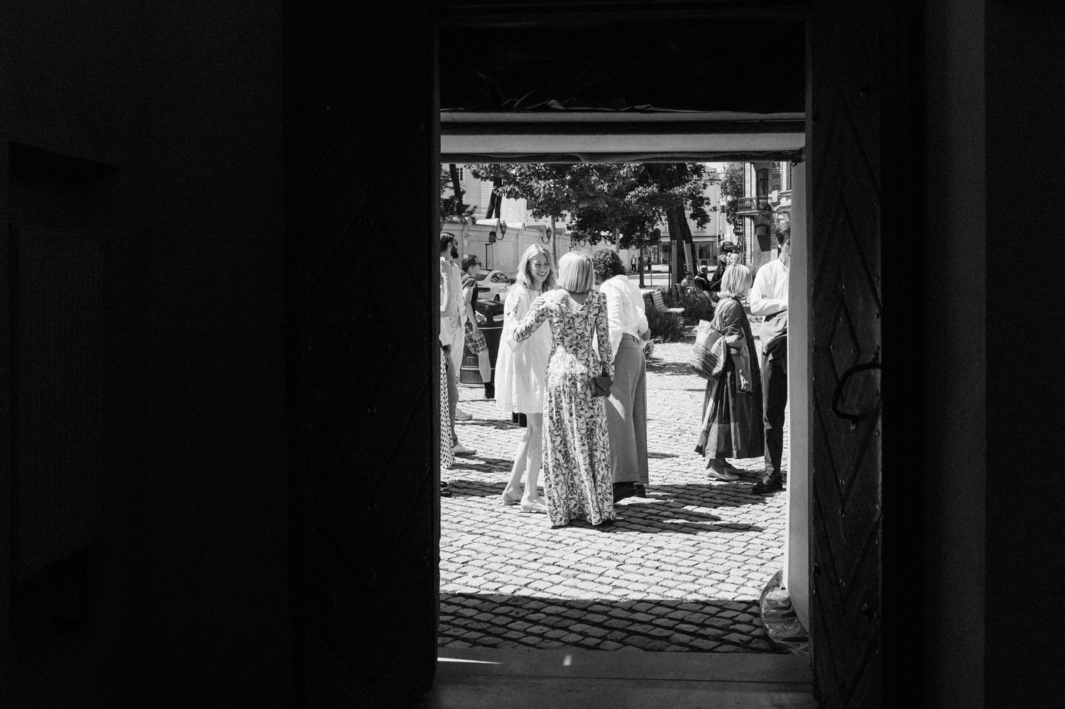 People in formal attire gather on a cobblestone courtyard viewed through a dark doorway.
