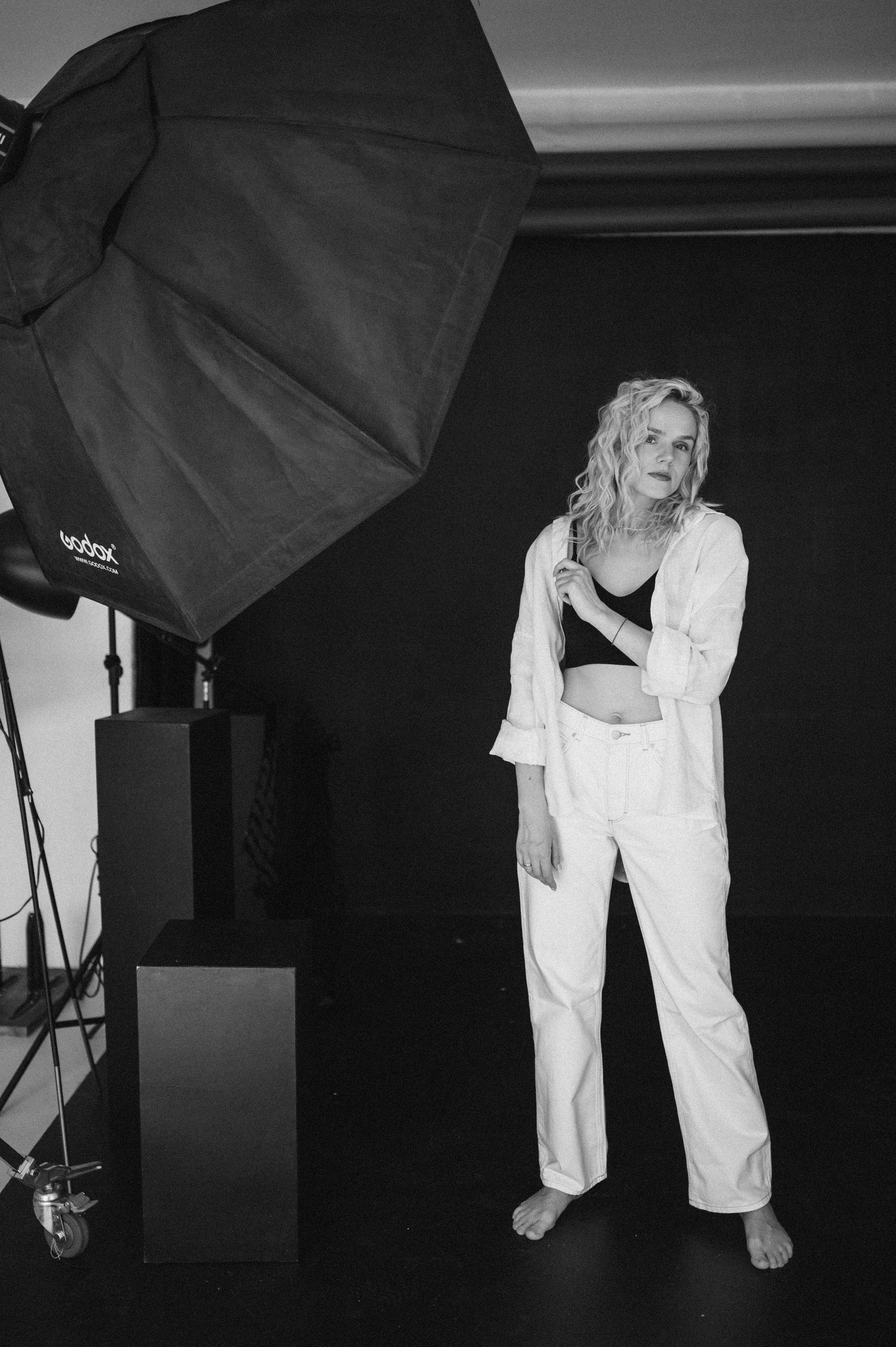 Black and white photo of a blonde woman in white pants and crop top posing in a photography studio with softbox lighting.