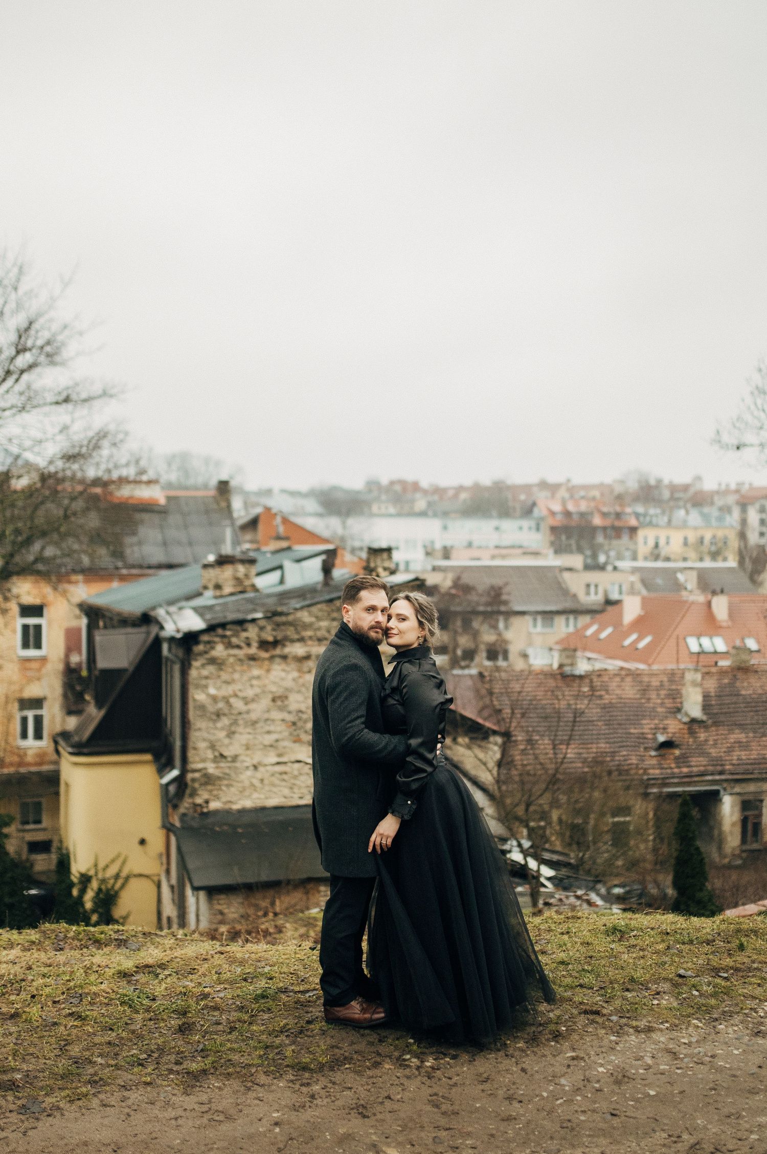 Couple sitting on hillside overlooking urban landscape with old buildings in background.