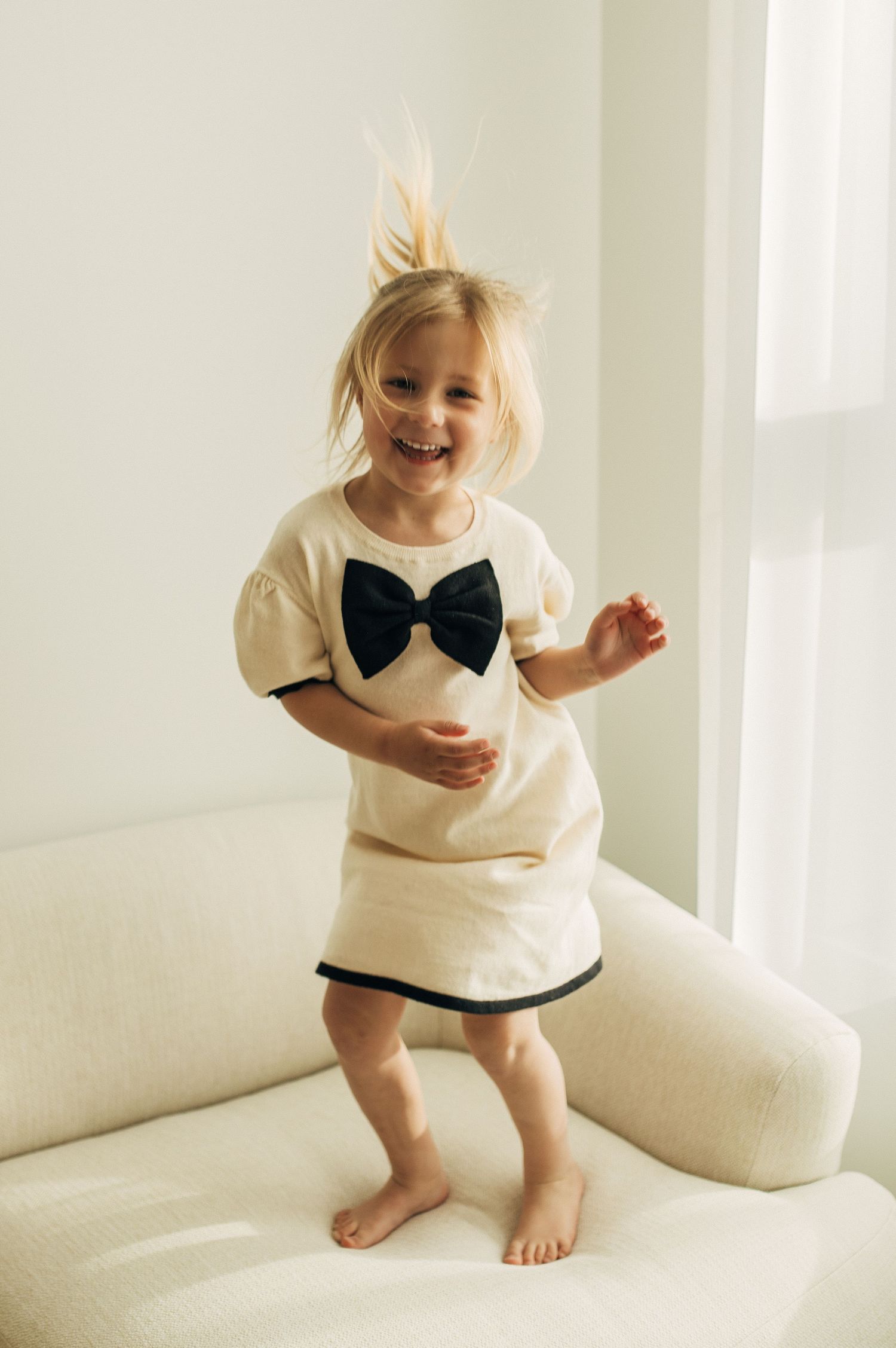 Small child wearing a white dress with black bow jumps playfully on white couch in bright room.