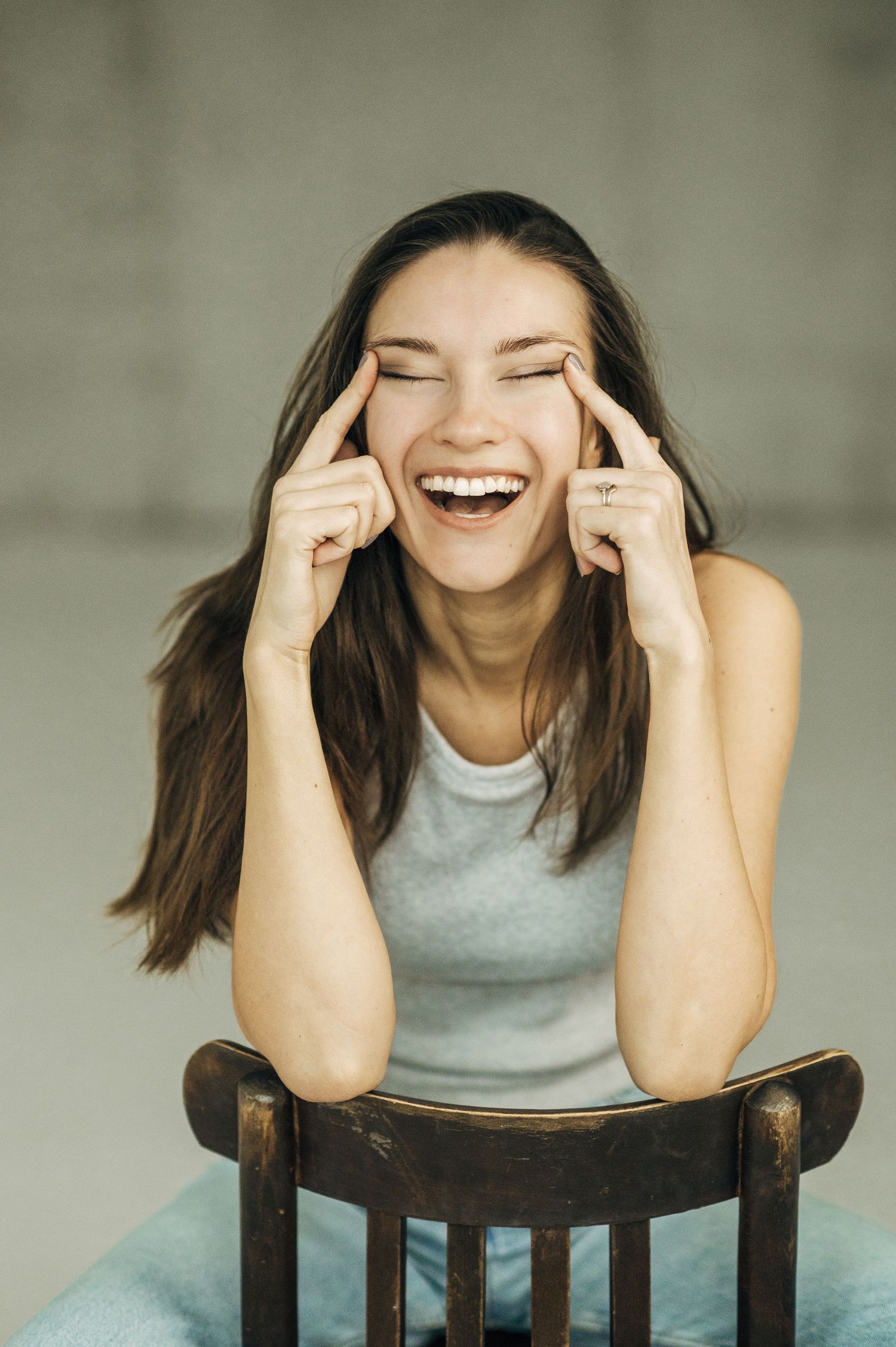 Joyful portrait of someone laughing while sitting on wooden chair in casual attire.