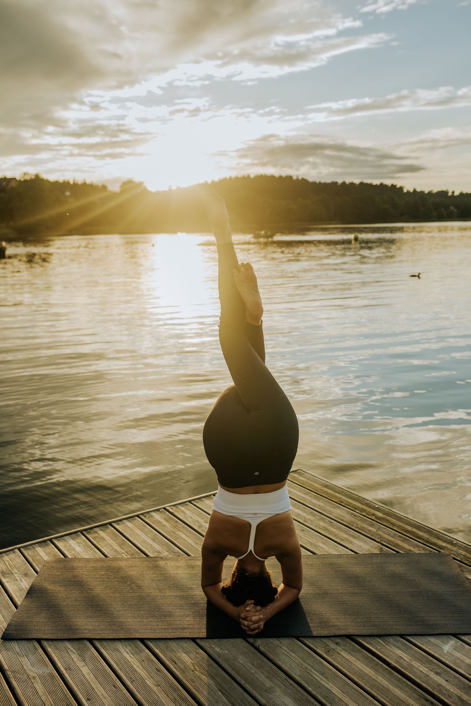 Silhouetted person practices yoga headstand on lakeside dock at sunset.