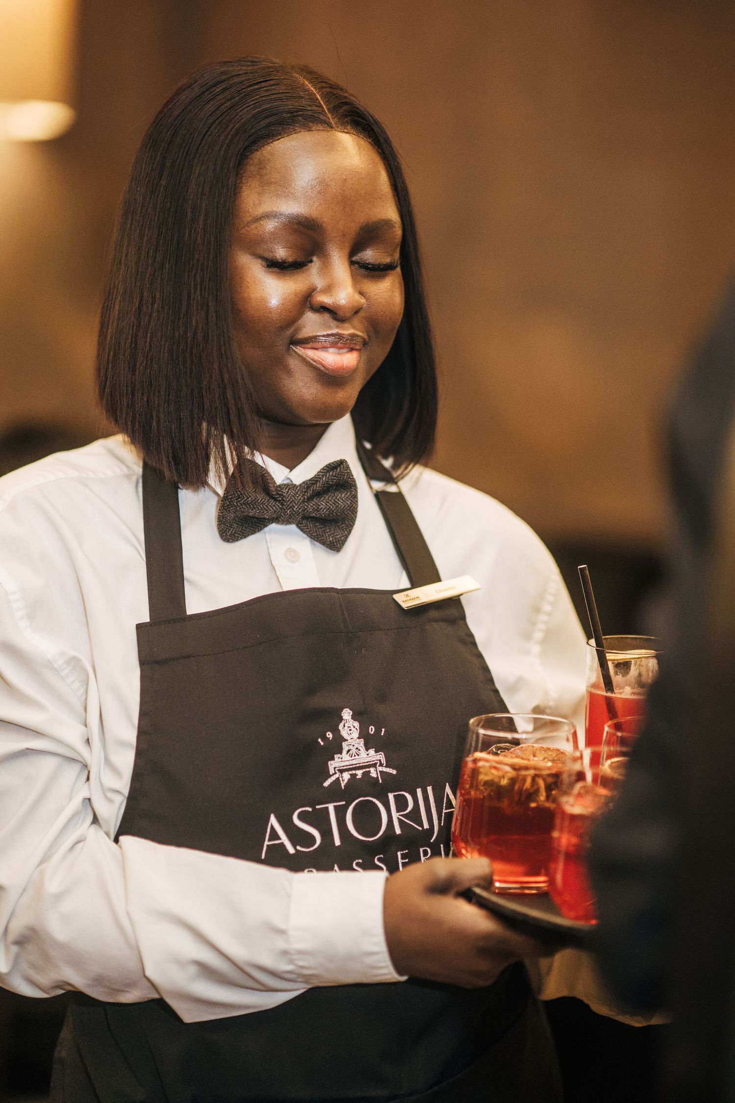 Server in white shirt and black apron smiles while holding a red cocktail at Astoria Bar and Lounge.