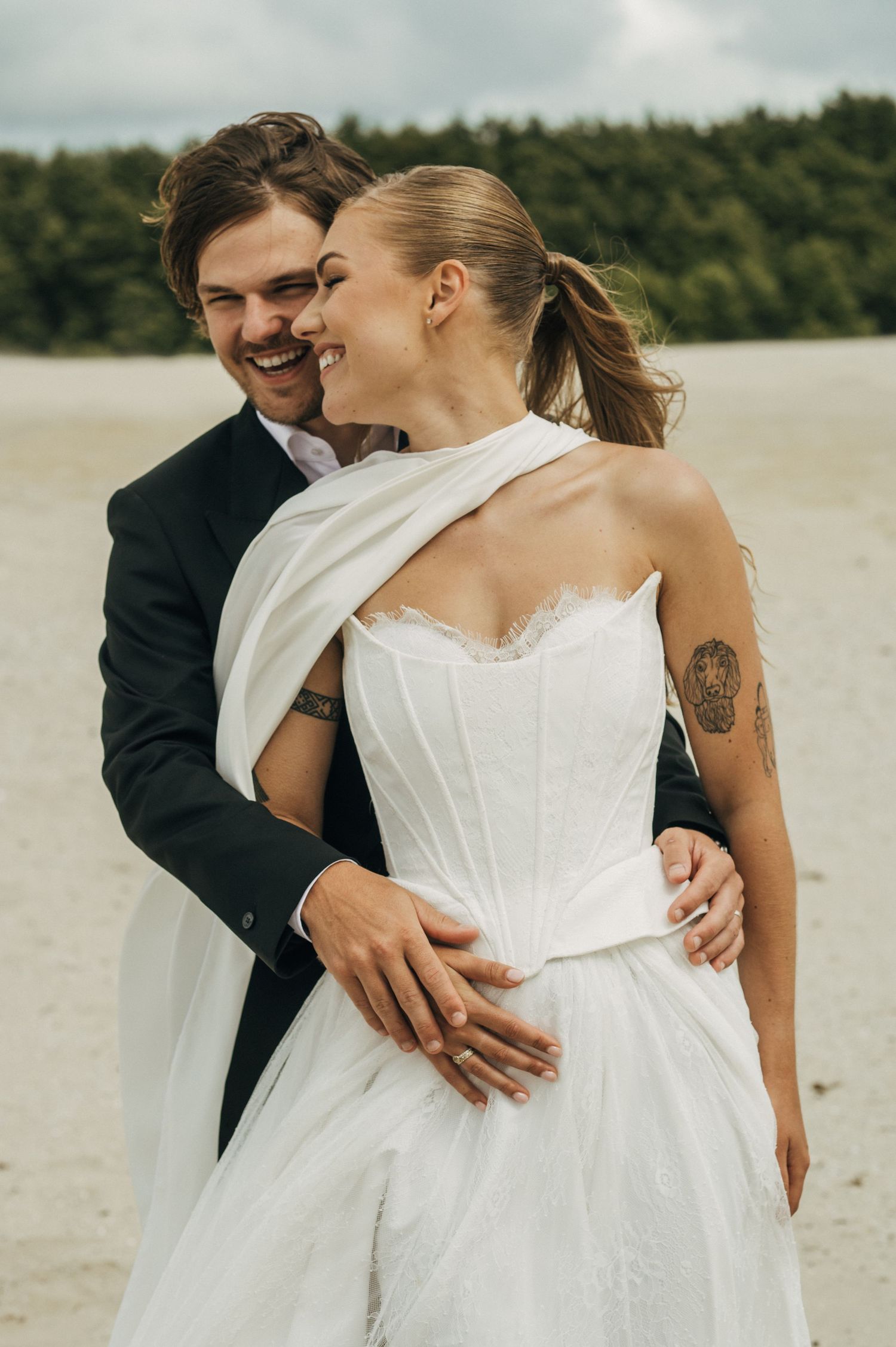 Bride and groom embrace on sandy beach, laughing joyfully, bride in strapless white gown with wrap, groom in dark suit.