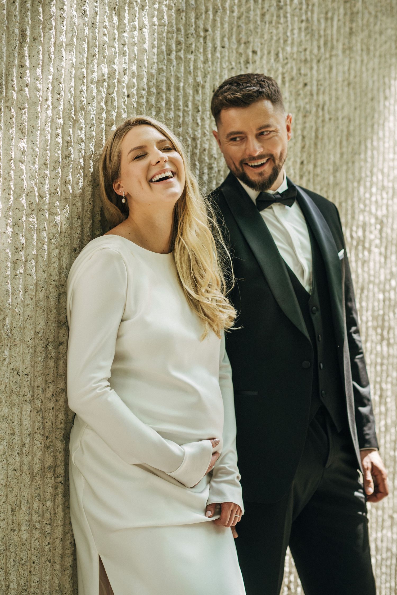 Smiling bride in white long-sleeve gown and groom in black tuxedo posing together against a shimmering wall.