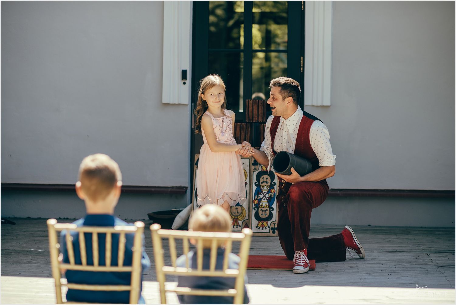 Magician entertains a young girl at an outdoor wedding reception while guests watch from gold chairs.