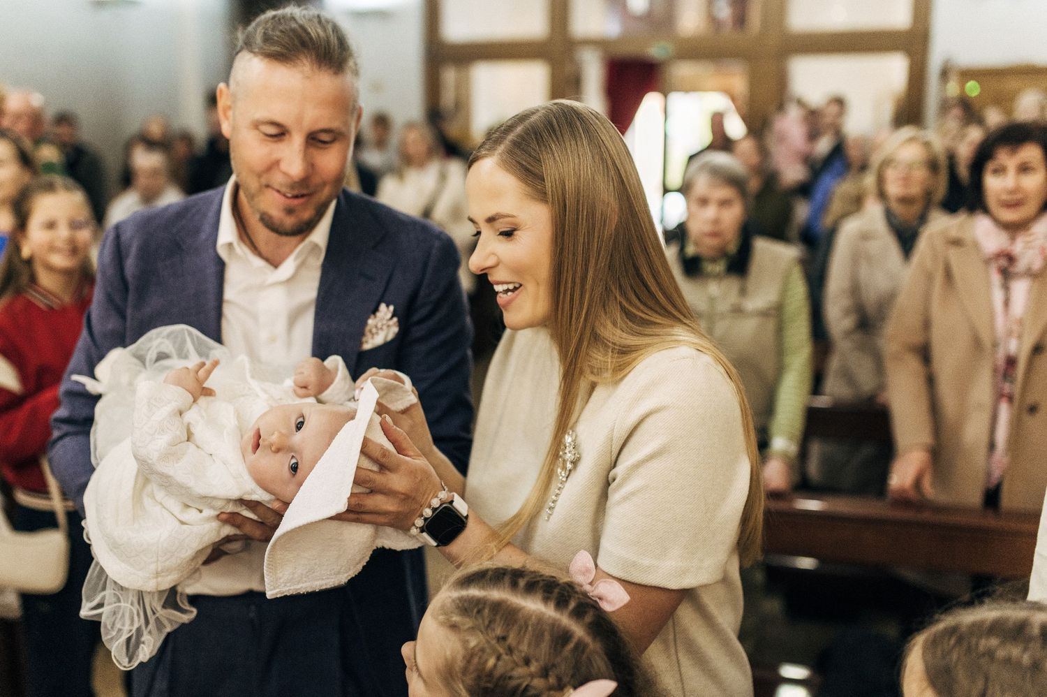 Parents holding newborn baby in white christening gown at a church baptism ceremony.