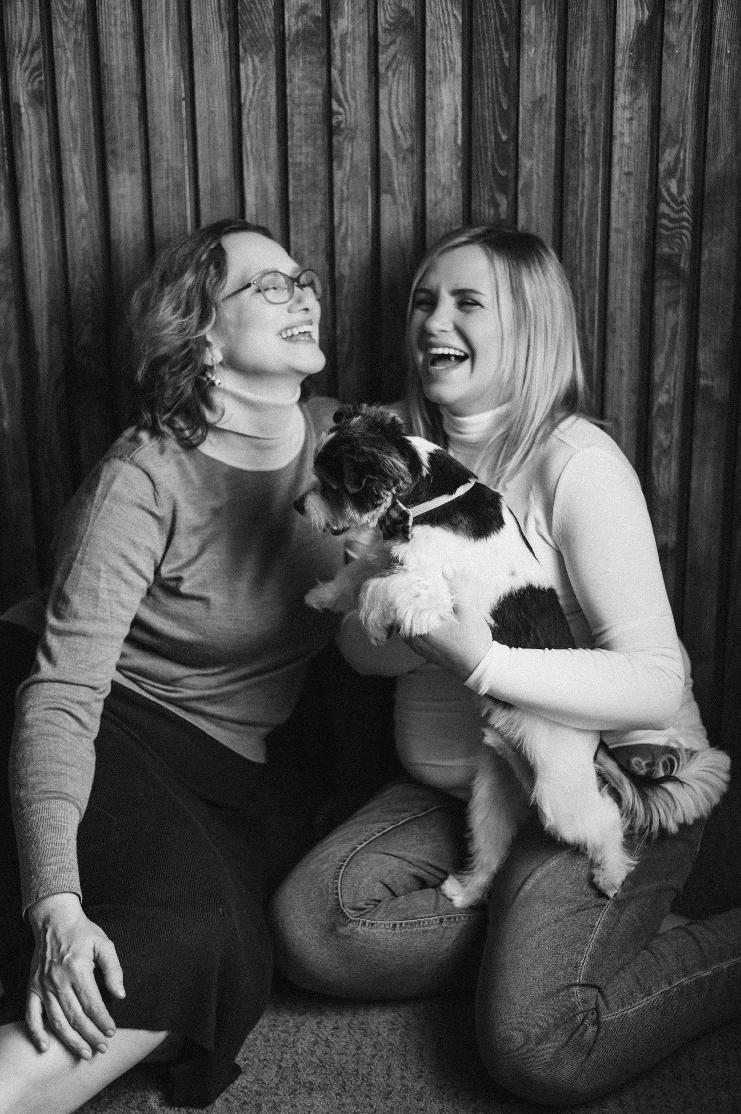 A black and white photo shows a group lounging casually with their pet dog against a wooden wall.