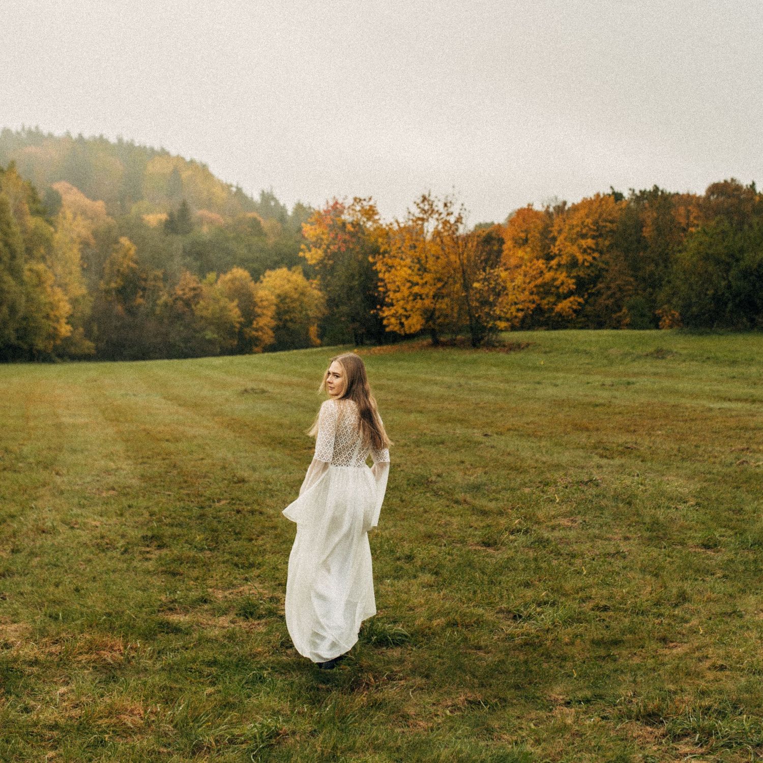 A figure in a flowing white dress walks through a misty autumn meadow with golden trees in the background.