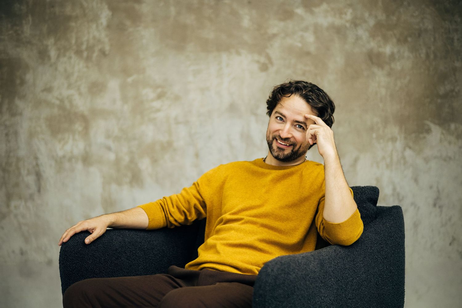 Bearded man in yellow sweater sitting relaxed on gray sofa against textured wall.
