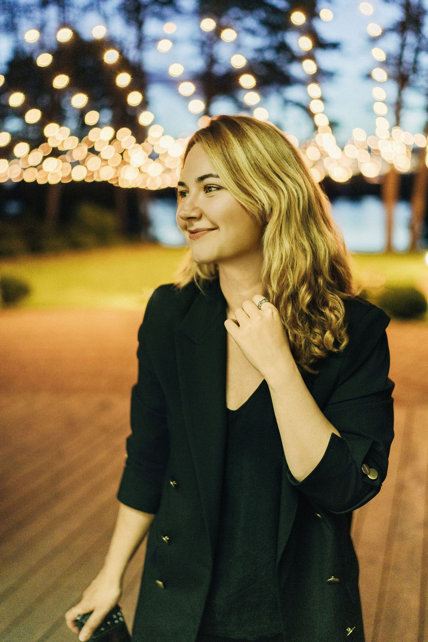 Woman in black blazer poses against twinkling lights in outdoor evening setting.