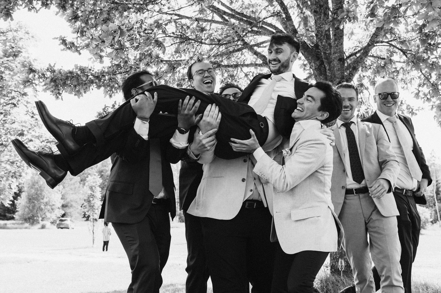 Wedding party poses playfully outdoors under trees in black and white.