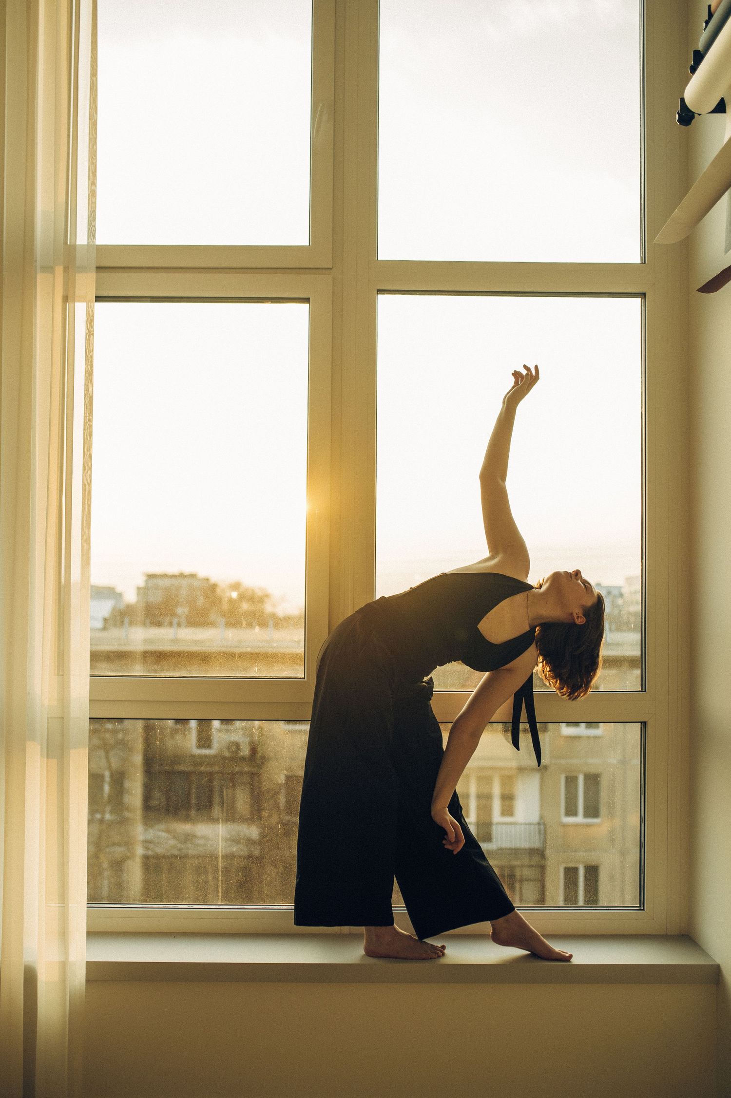 A graceful dancer in black outfit poses elegantly on a windowsill, backlit by golden sunset light.