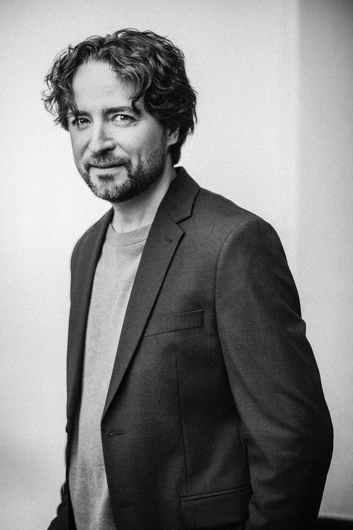 Black and white portrait of bearded man in blazer standing against light background.