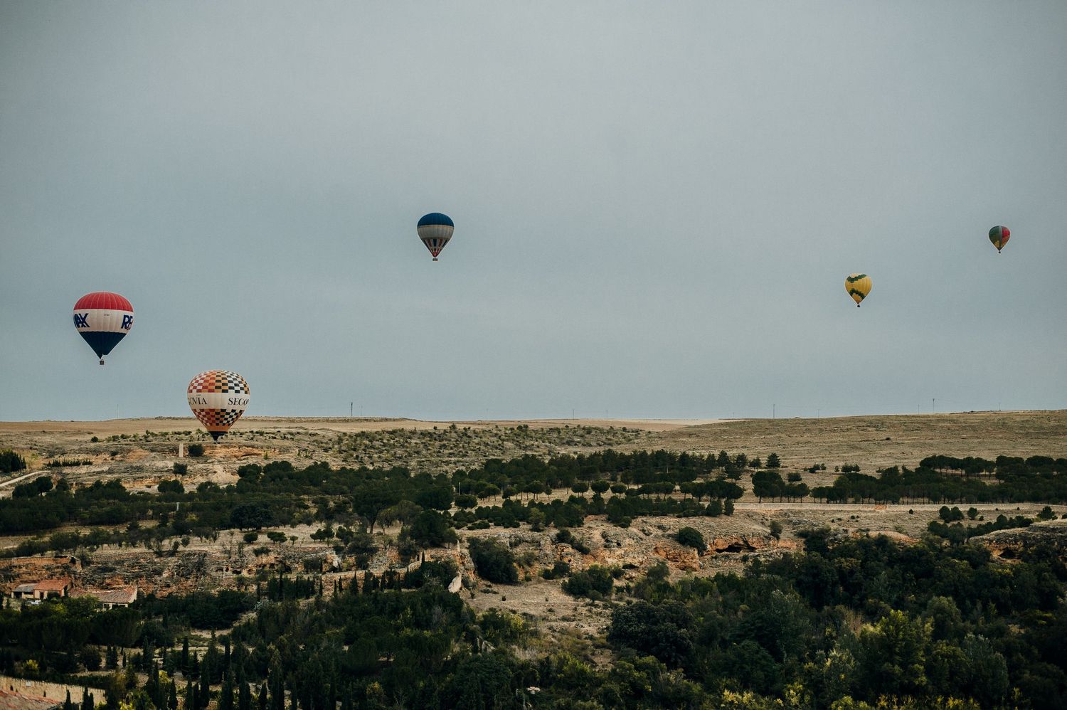 Hot air balloons float over a desert landscape with scattered buildings and vegetation during golden hour.