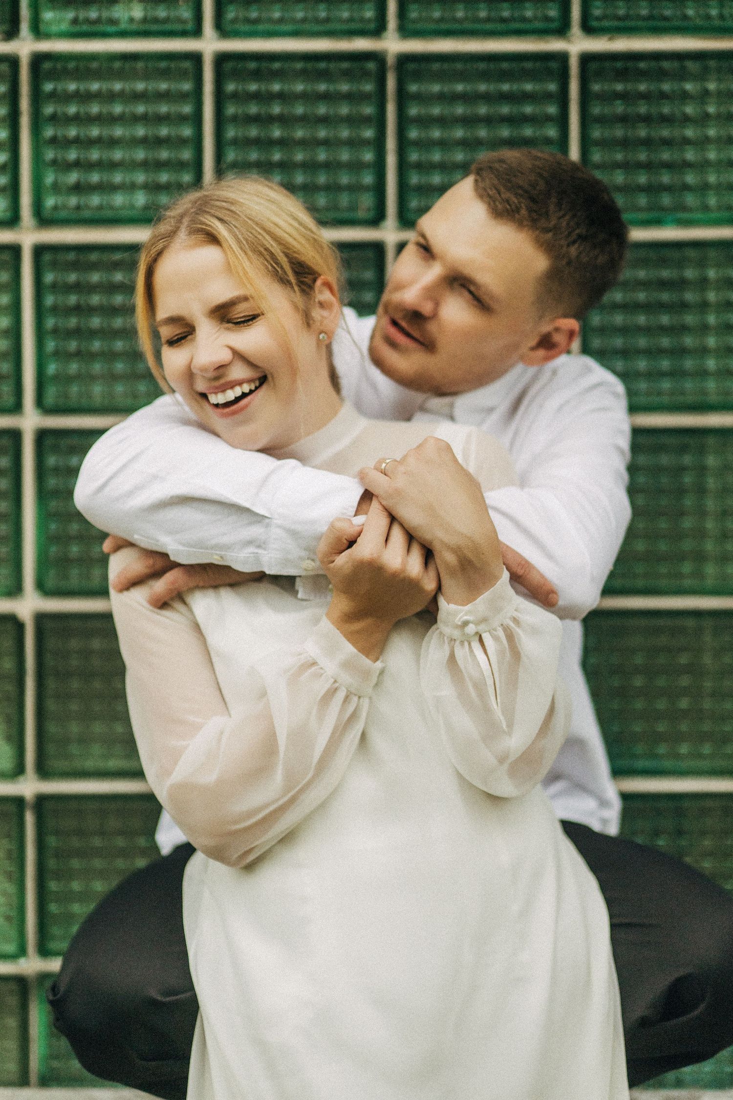 Two people in white shirts embrace and laugh together against a green tiled wall background.