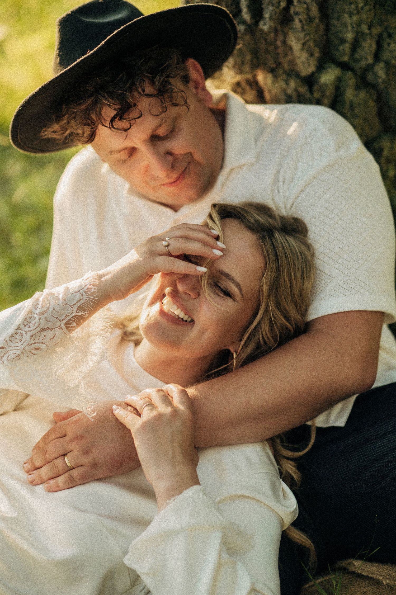 A romantic couple embraces outdoors in sunlight, with one wearing a black hat and white shirt while the other laughs joyfully.