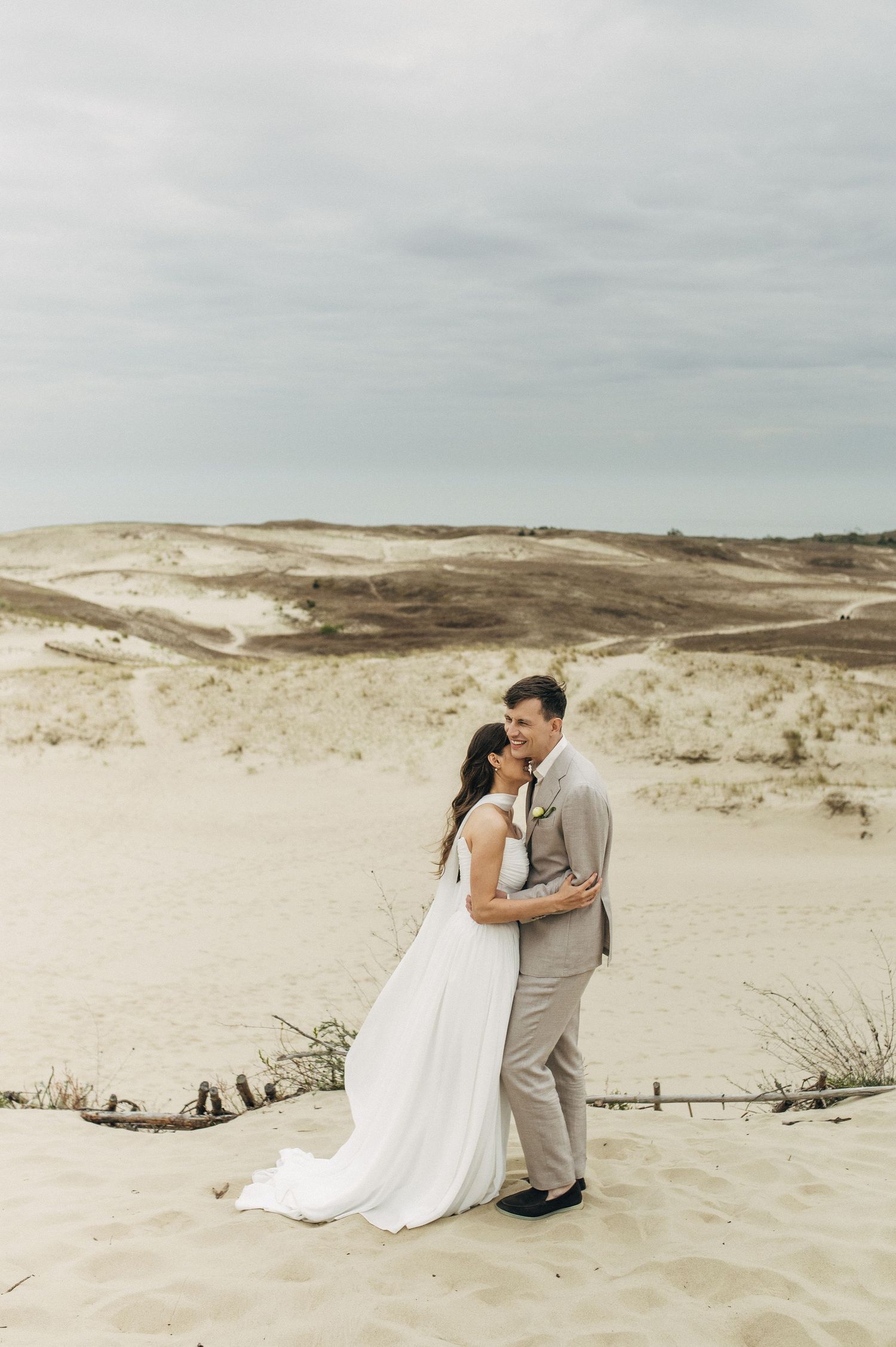 A romantic couple embraces on sandy dunes during a beach photoshoot wearing white and gray formal attire.