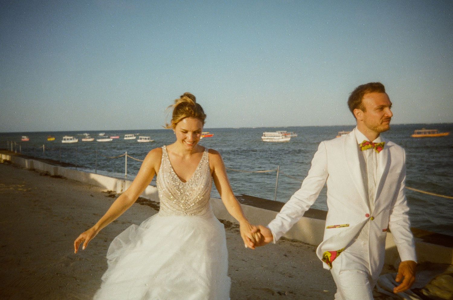 A couple in wedding attire walks hand in hand along a scenic beach at sunset with boats in the background.