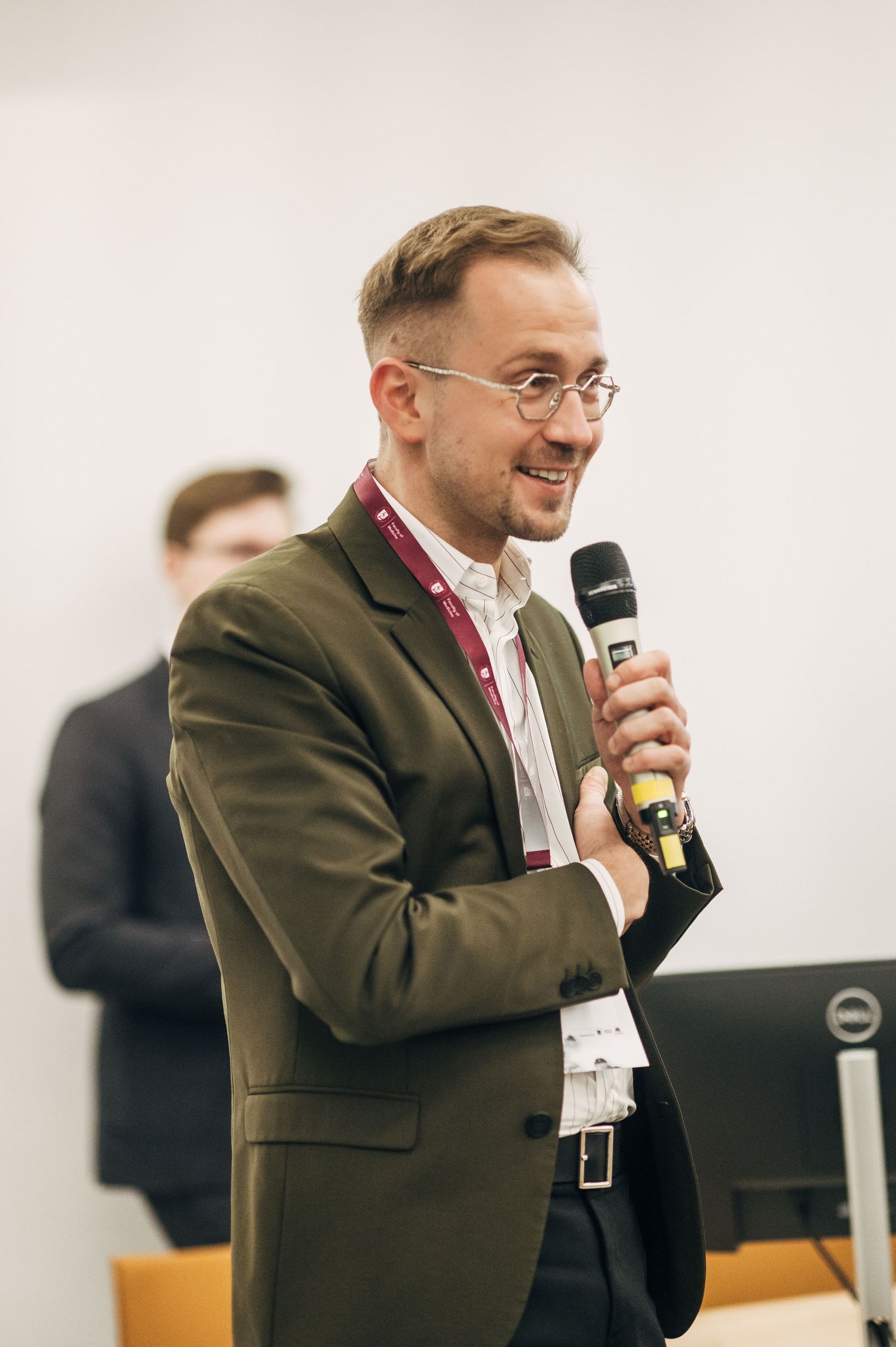 Speaker in brown jacket and glasses smiles while presenting at a professional event with microphone in hand.