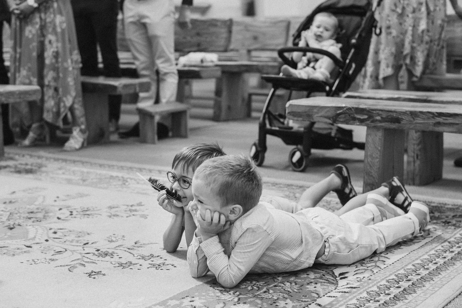 Two children lying on a rug playing while a baby sits in a stroller at a family gathering.