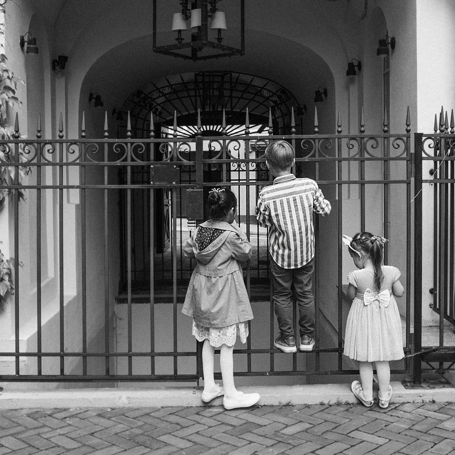 Three children stand at ornate iron gates of an elegant estate, viewed from behind in black and white.