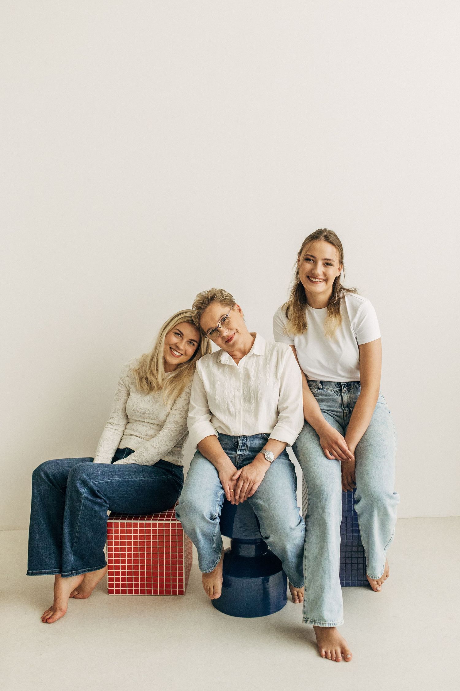 Three smiling women in casual jeans and white tops sitting together on colorful cube seats against a white wall.