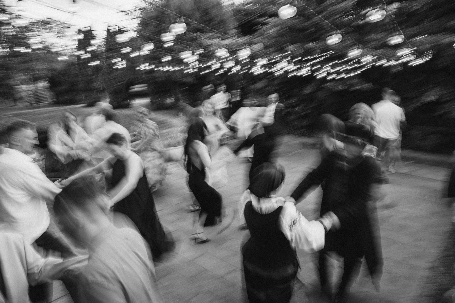 Black and white photo of couples dancing outdoors at a wedding reception under string lights.