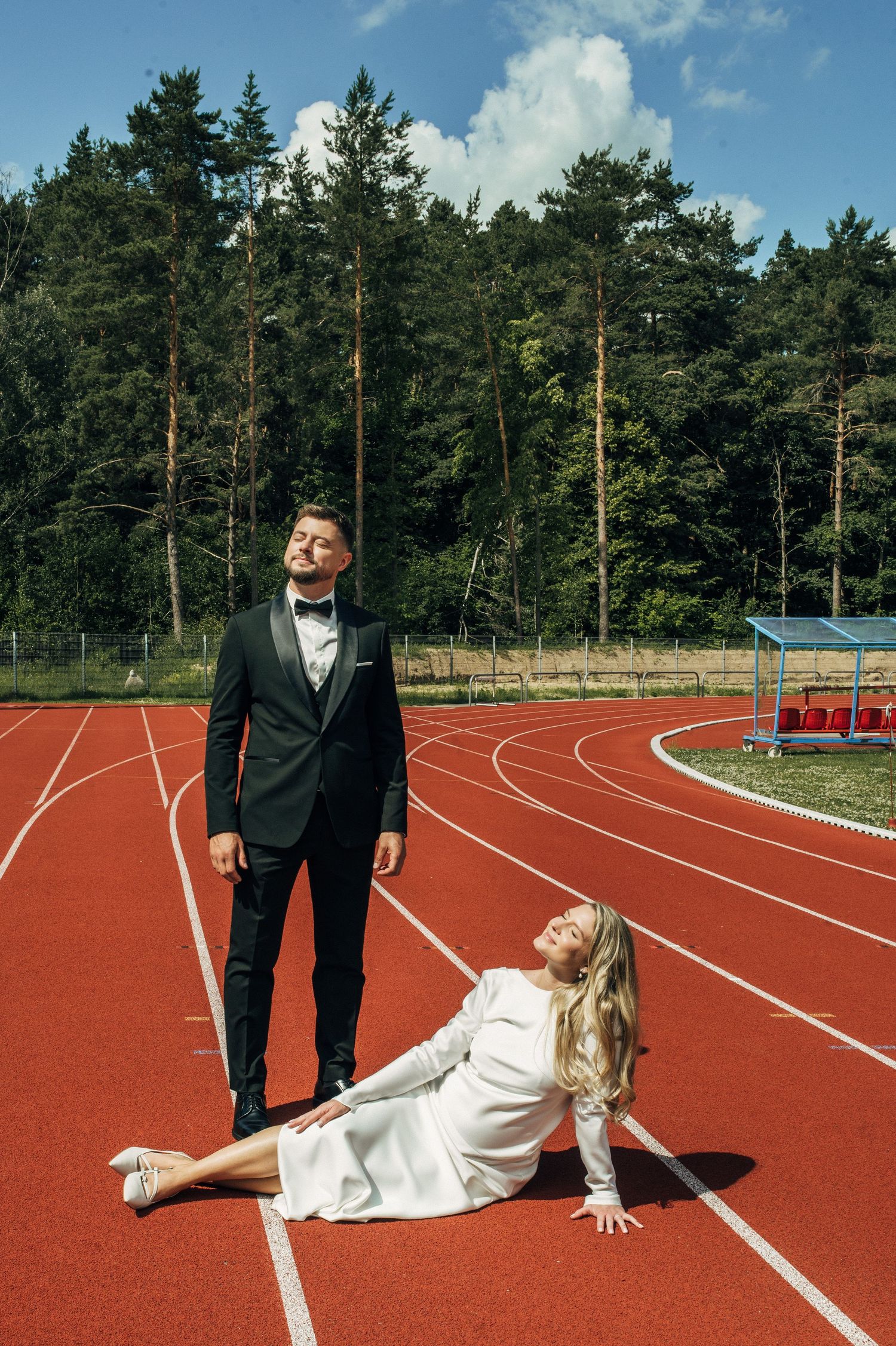 Bride in white dress sits on red running track while groom in black tuxedo stands beside her outdoors.