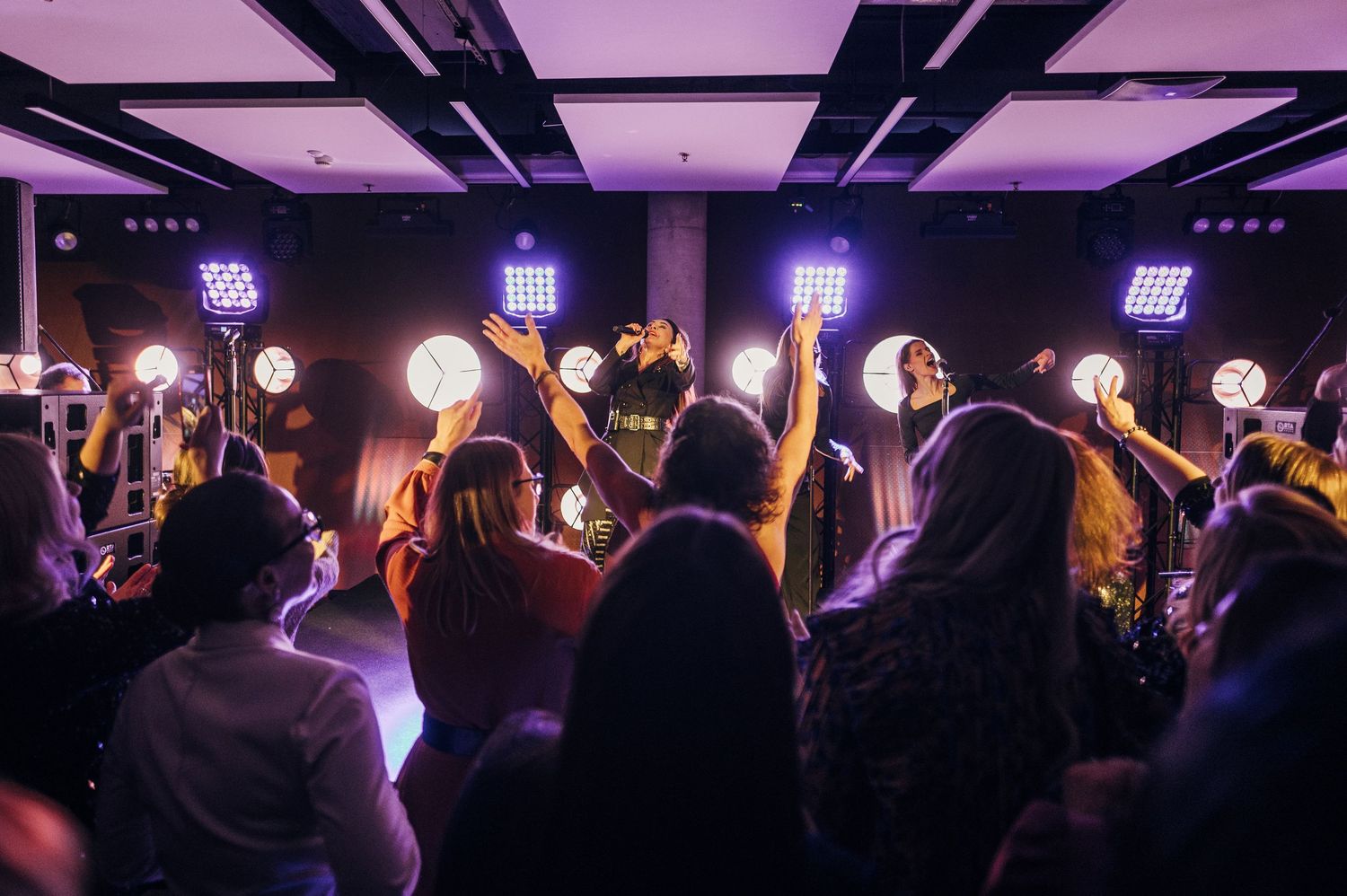 Concert crowd with raised hands watching performers on stage with bright illuminated circular lights in dark venue.