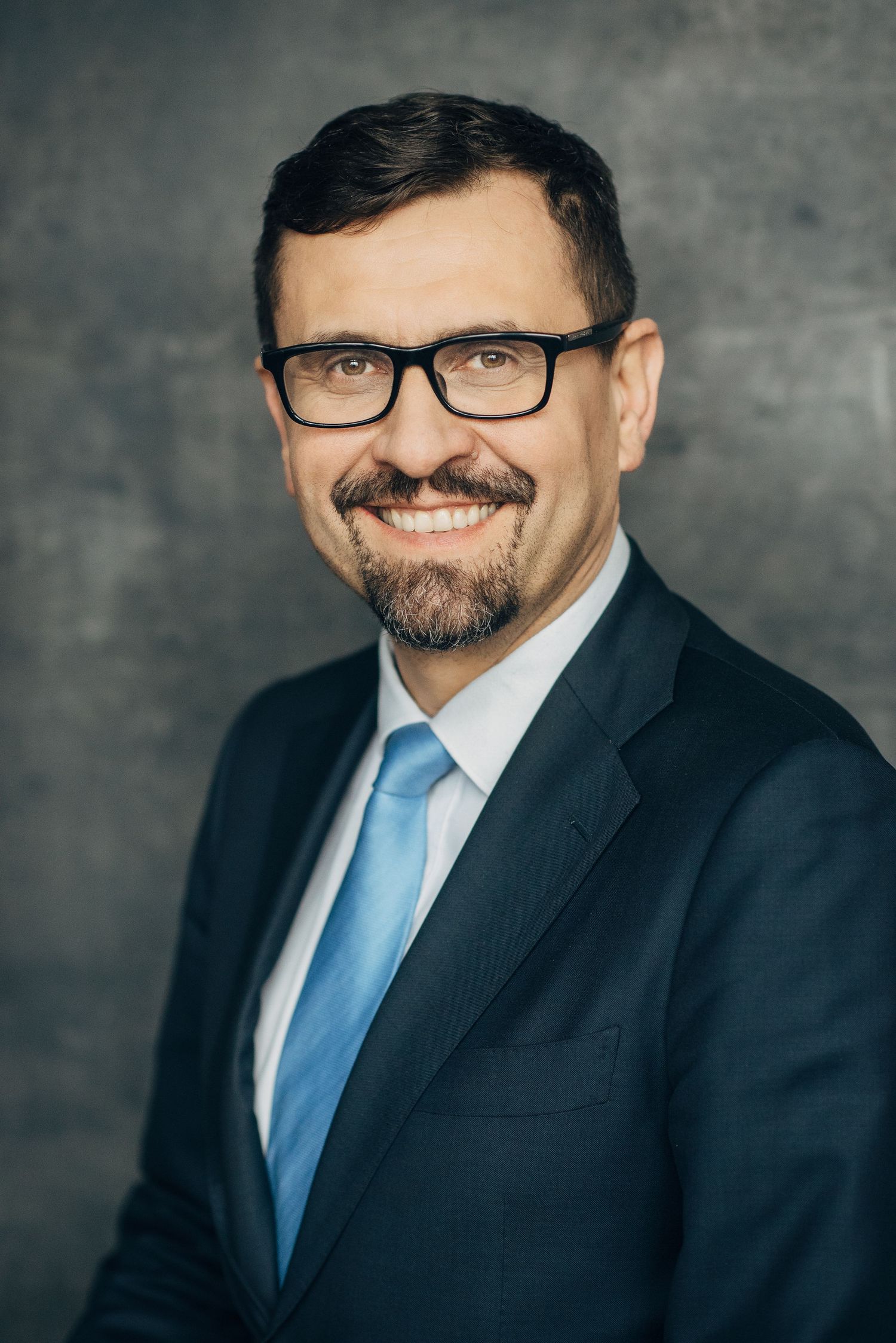 Business portrait of a person wearing glasses, blue tie and dark suit against textured background.