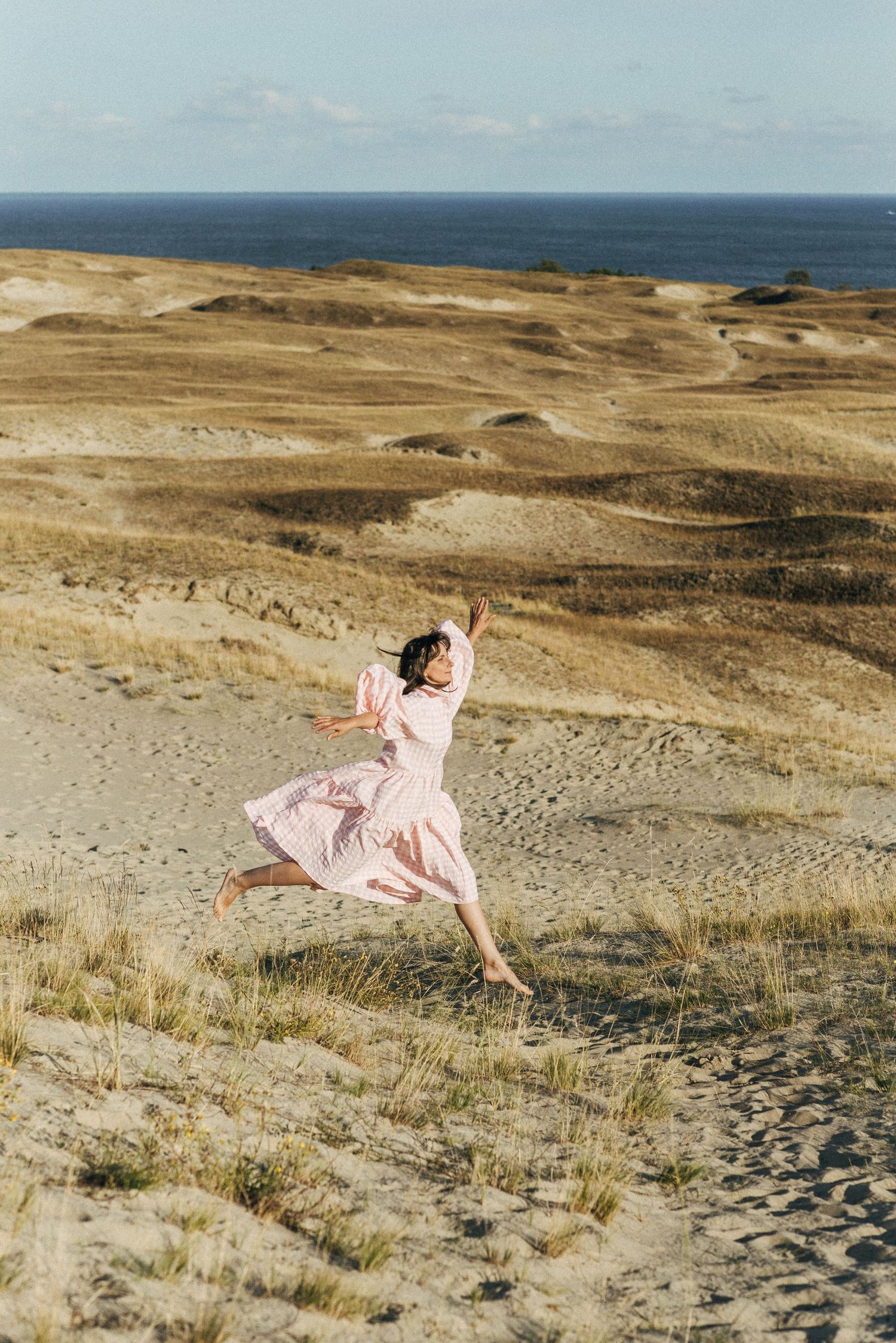 Person in pink dress jumping joyfully on sandy dunes with ocean view.