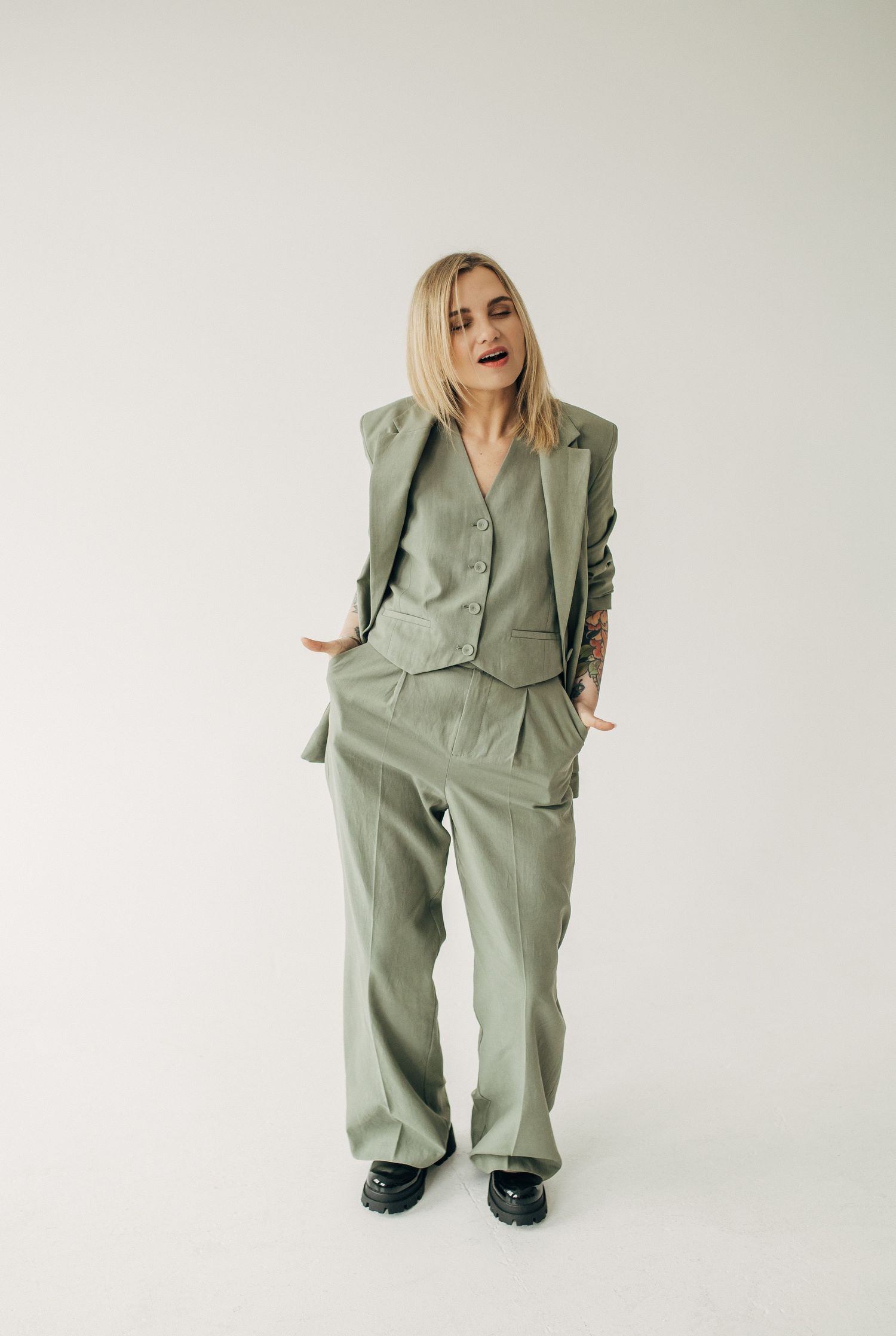Fashion model poses in oversized sage green suit and heels against white background in editorial series.