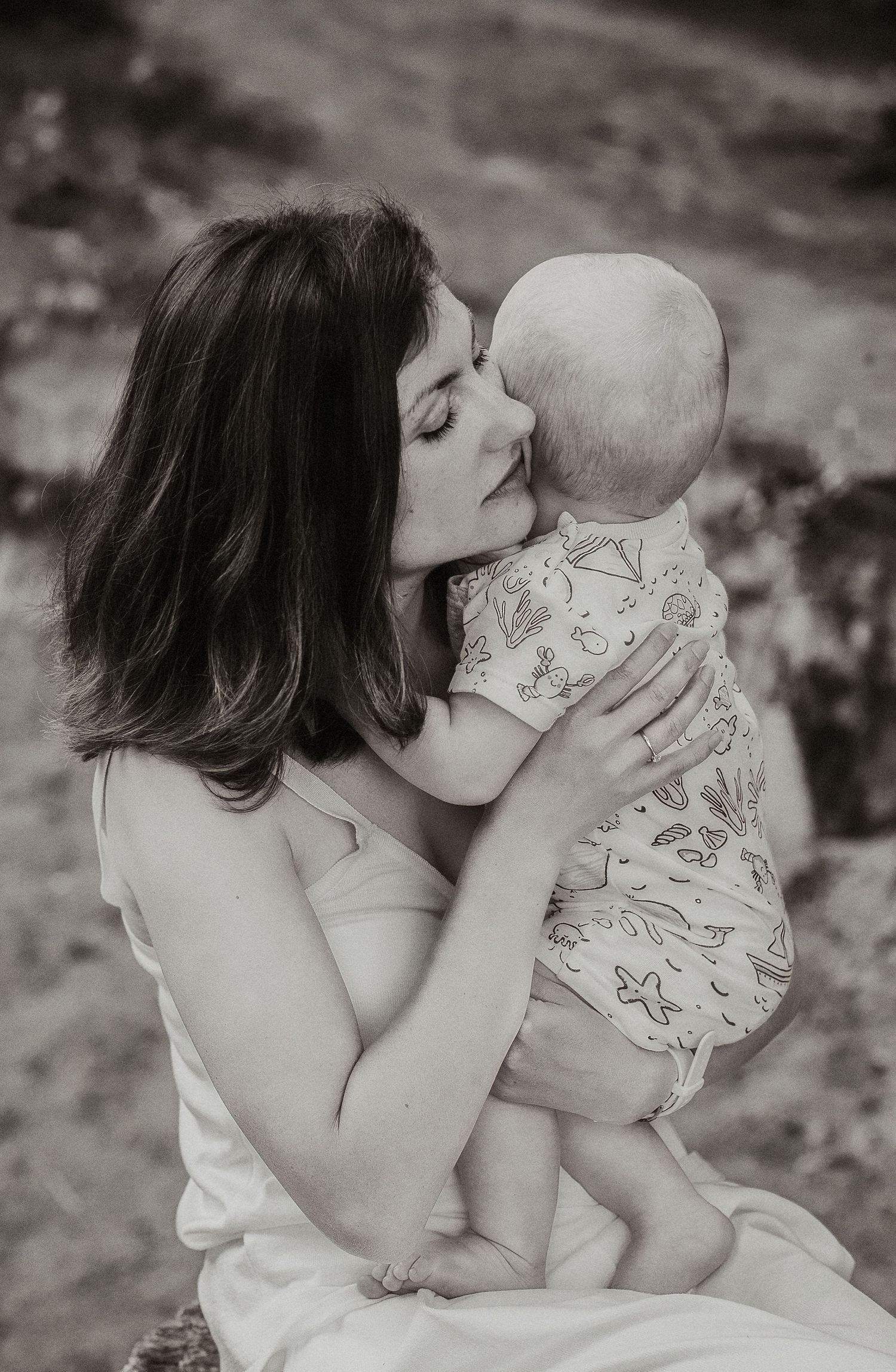 Black and white photo of a mother kissing her baby outdoors, capturing a tender moment of maternal love.