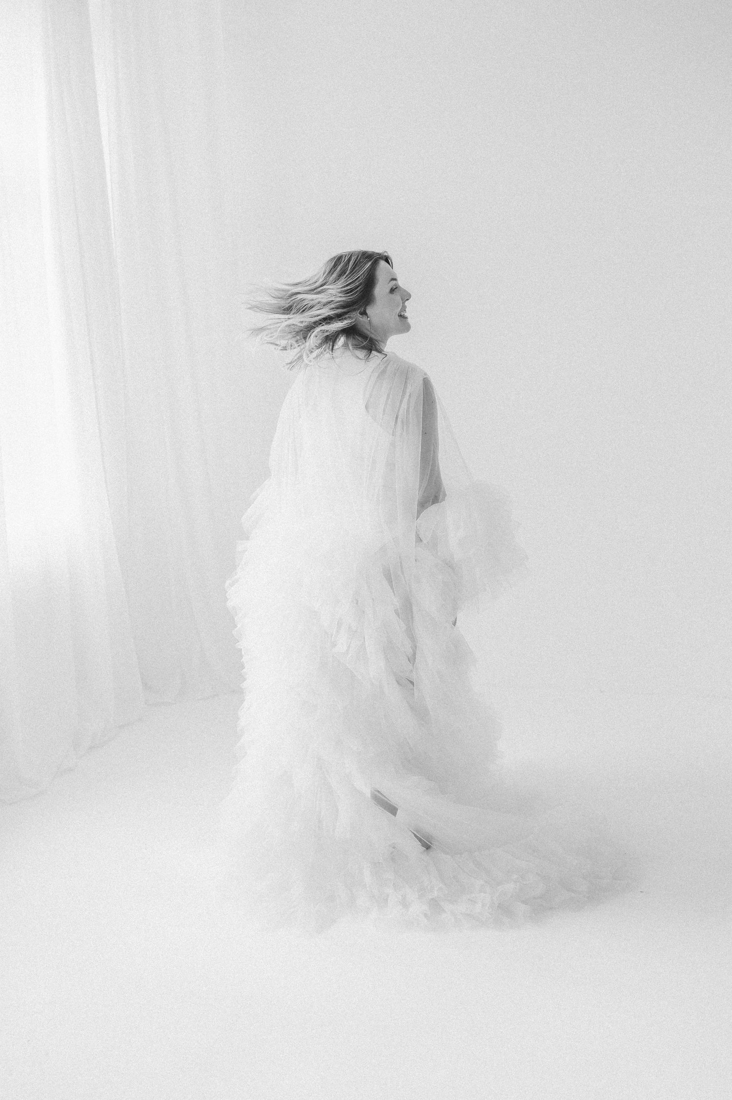 A woman in a flowing white ruffled gown twirls gracefully in a bright minimalist studio, captured in elegant black and white.