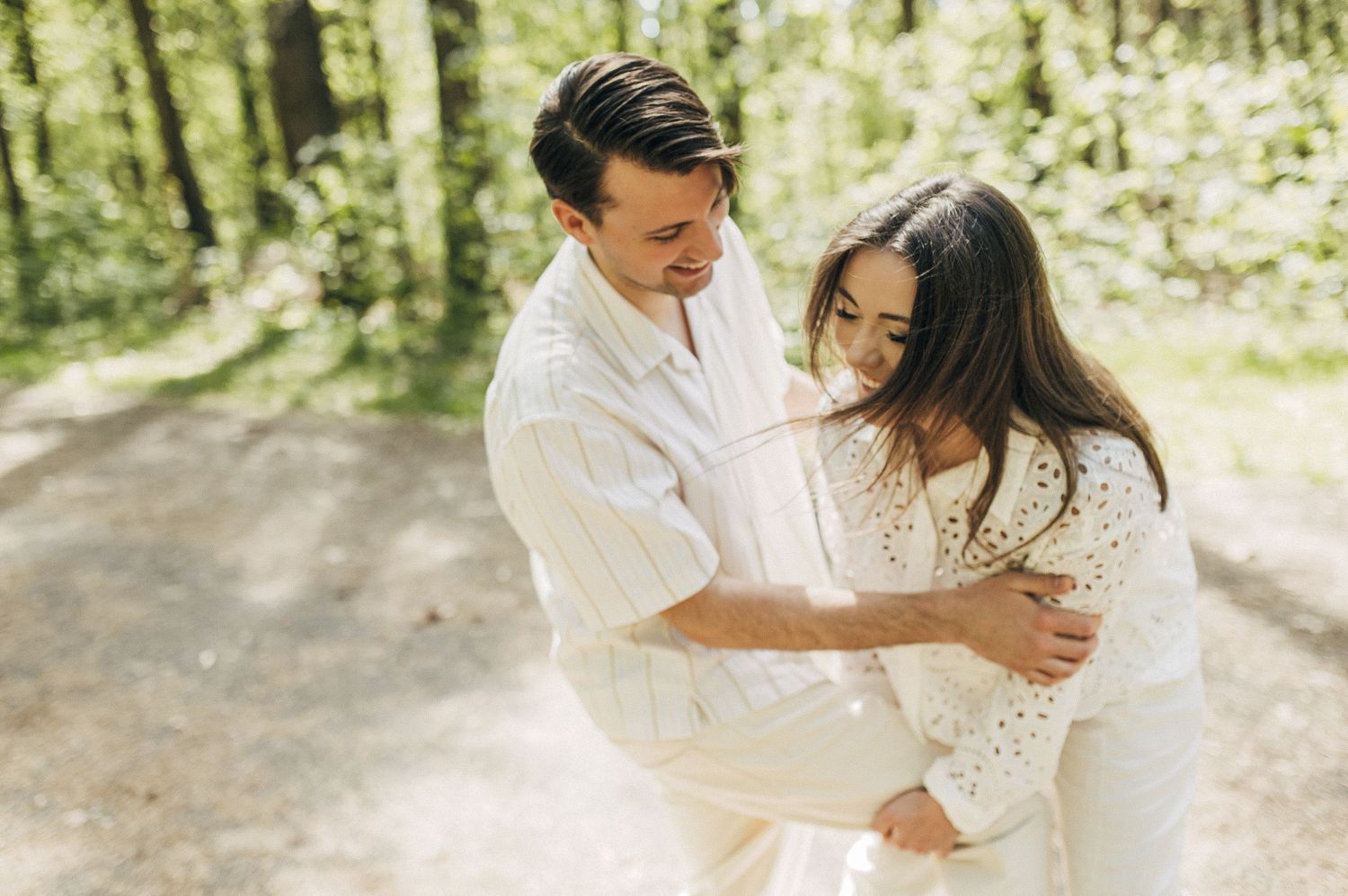 A joyful couple in white outfits sharing a playful moment on a sunny forest path.